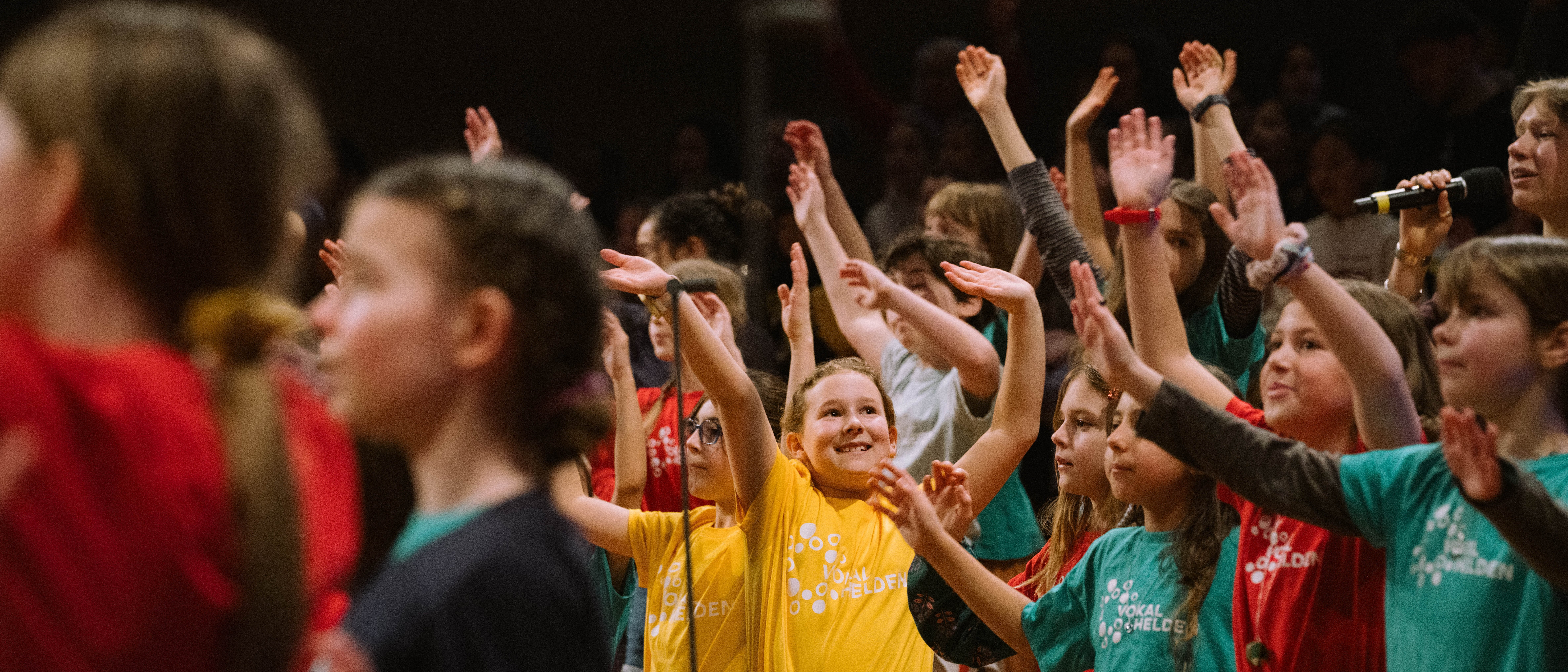 Eine Gruppe von Kindern in bunten T-Shirts hebt die Arme und lächelt, während sie gemeinsam auf der Bühne auftreten und singen.