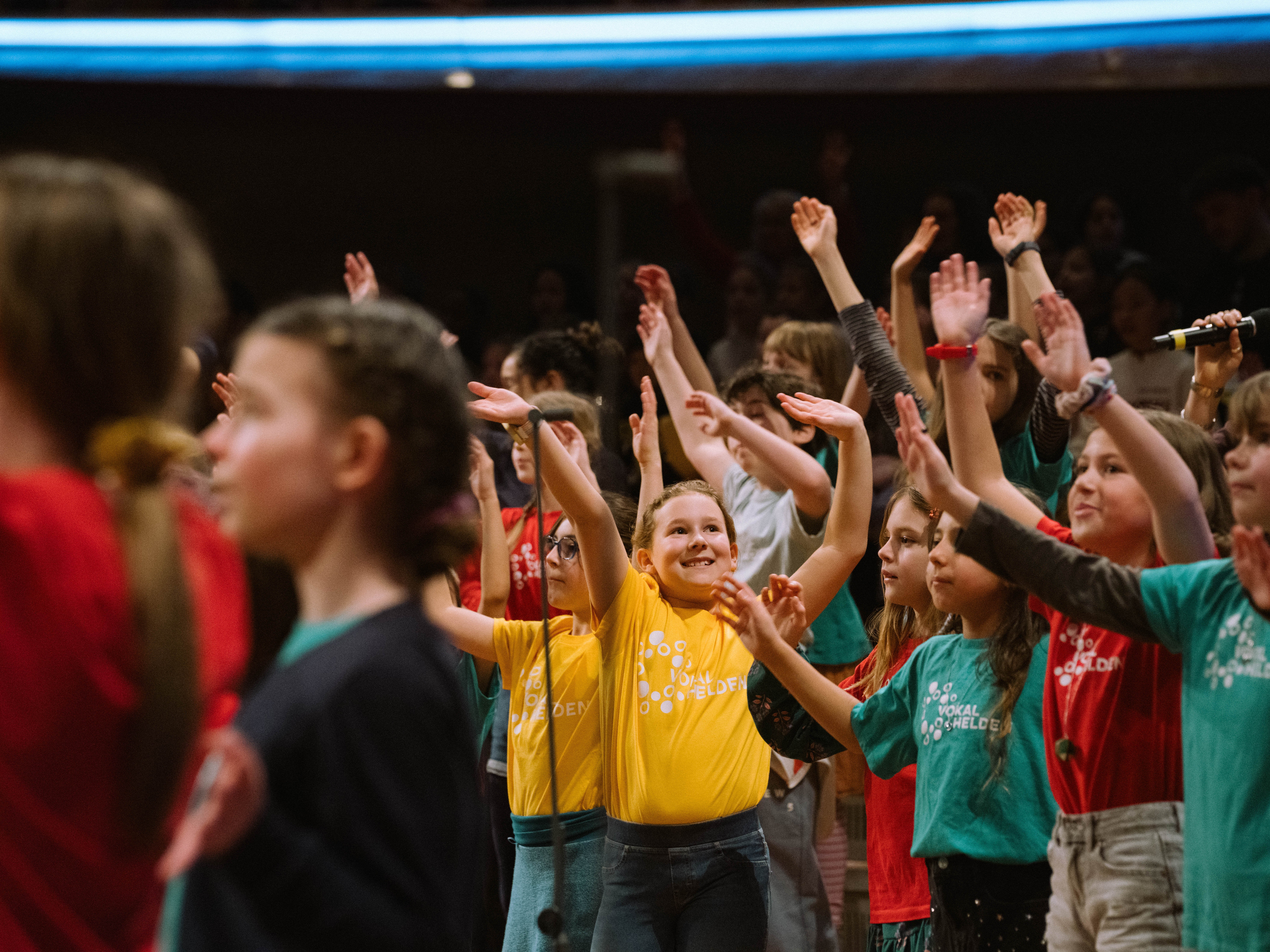 Eine Gruppe von Kindern in bunten T-Shirts hebt die Arme und lächelt, während sie gemeinsam auf der Bühne auftreten und singen.