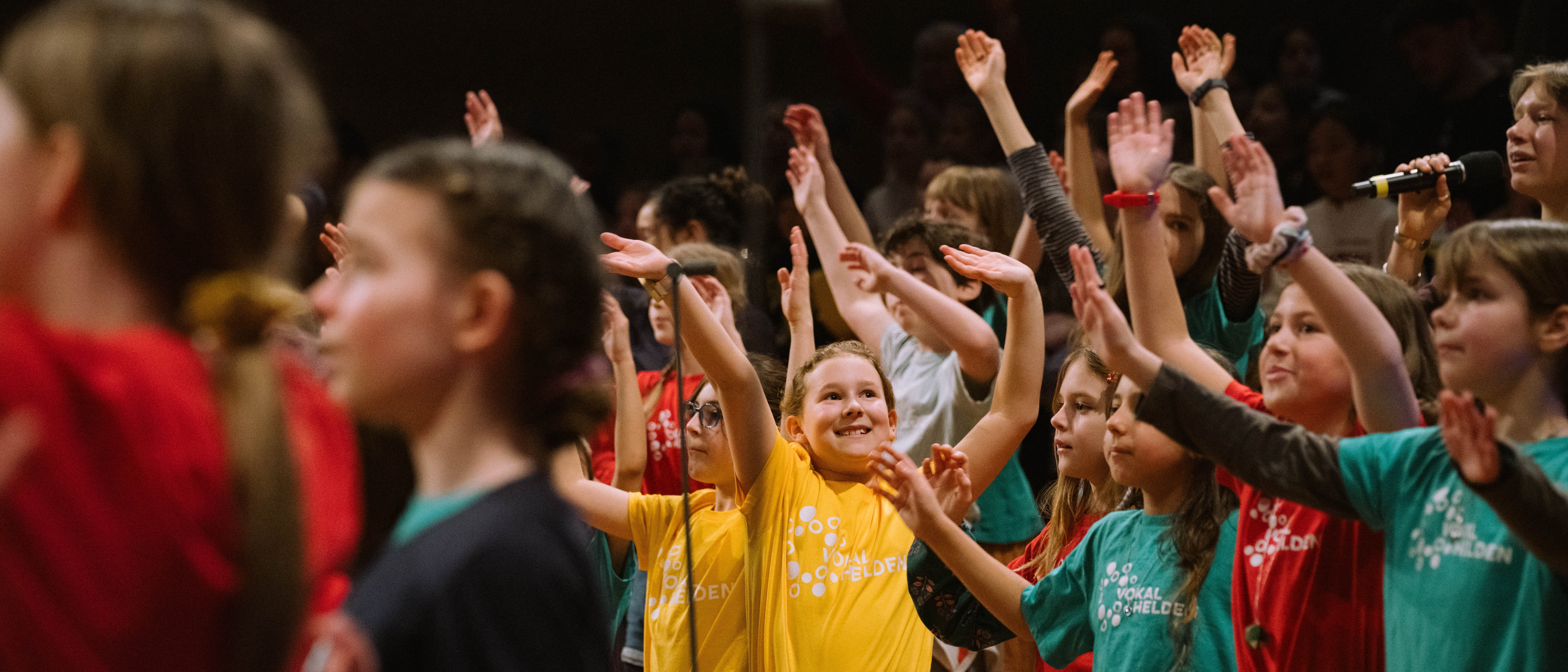 Eine Gruppe von Kindern in bunten T-Shirts hebt die Arme und lächelt, während sie gemeinsam auf der Bühne auftreten und singen.