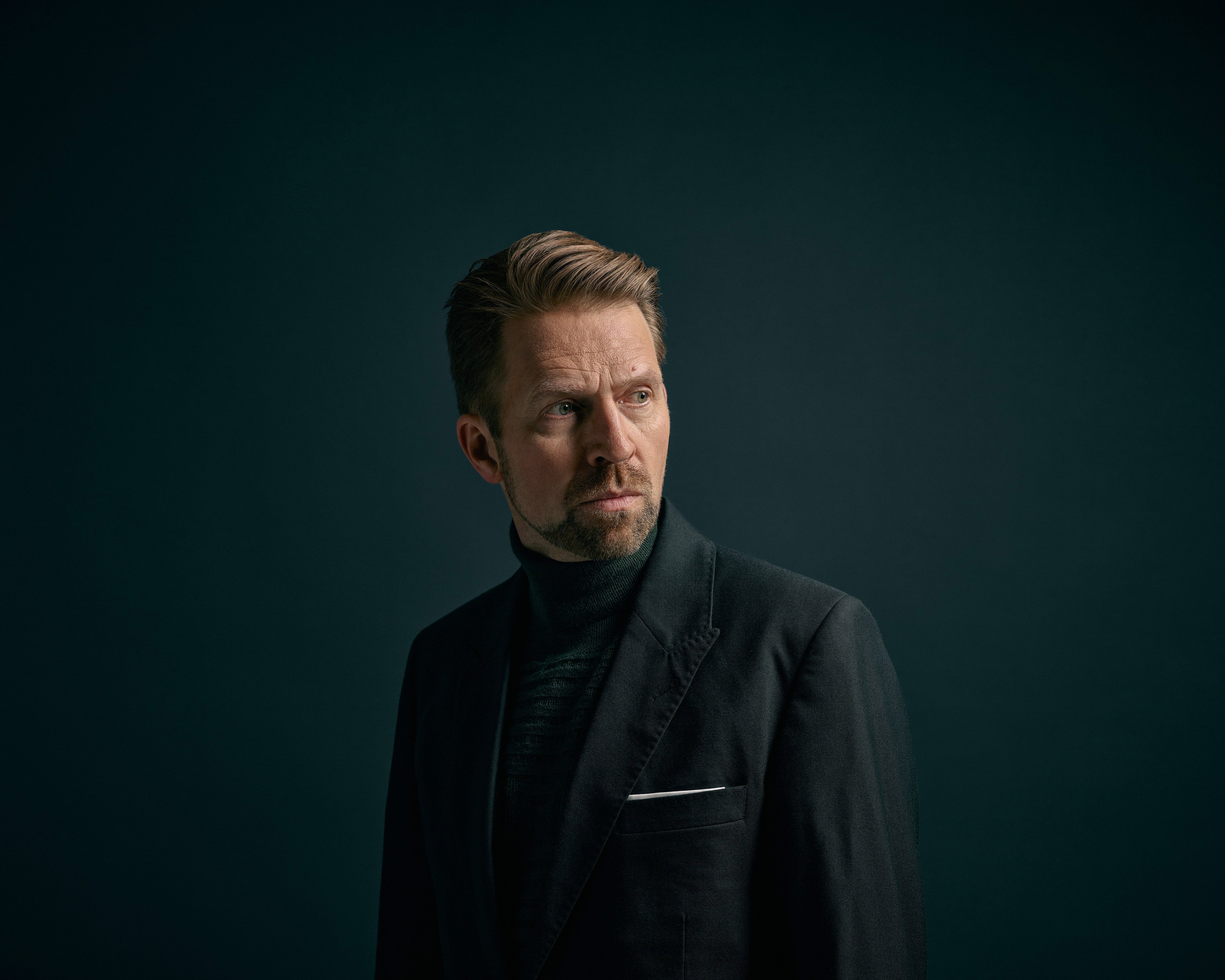 Leif-Ove Andsnes mit kurzem, hellbraunem Haar und gestutztem Bart, der einen dunklen Anzug und einen Rollkragenpullover trägt, steht vor einem dunklen Hintergrund und schaut nachdenklich nach links.
