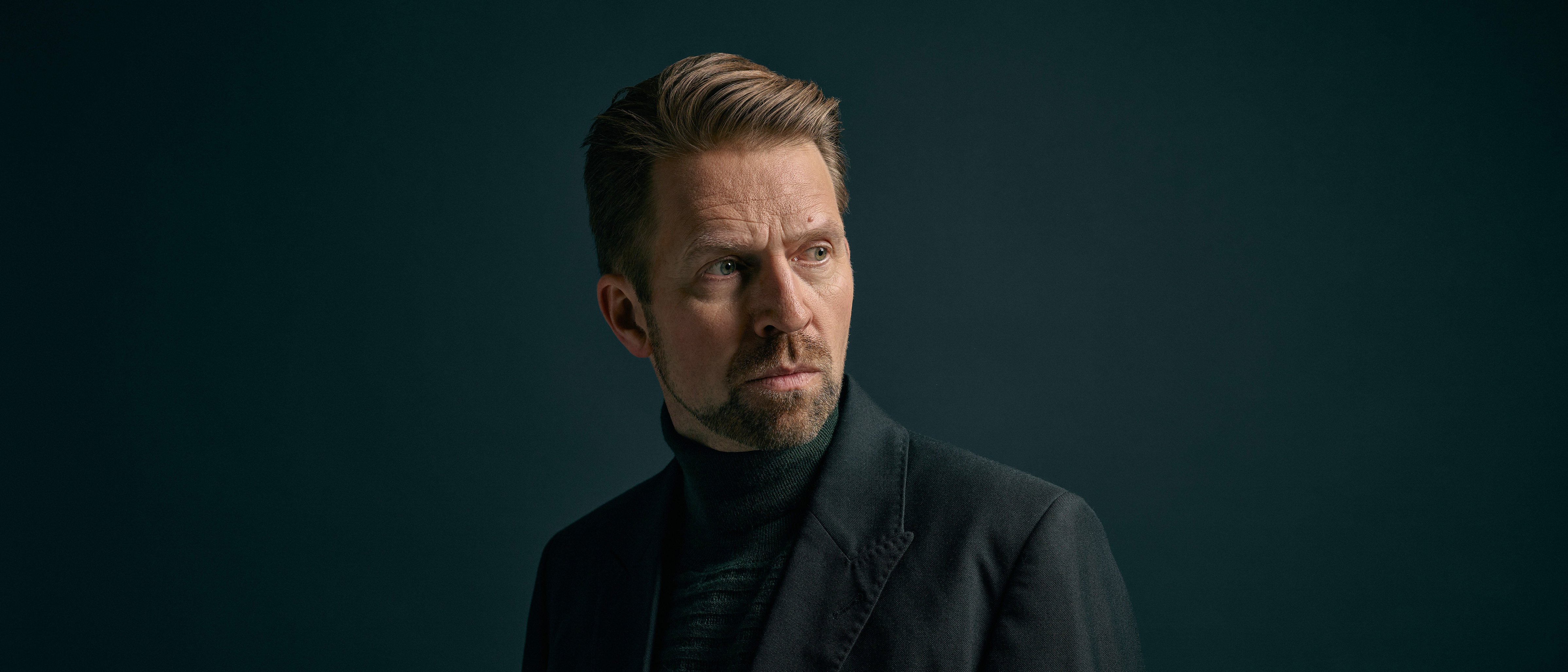 Leif-Ove Andsnes mit kurzem, hellbraunem Haar und gestutztem Bart, der einen dunklen Anzug und einen Rollkragenpullover trägt, steht vor einem dunklen Hintergrund und schaut nachdenklich nach links.