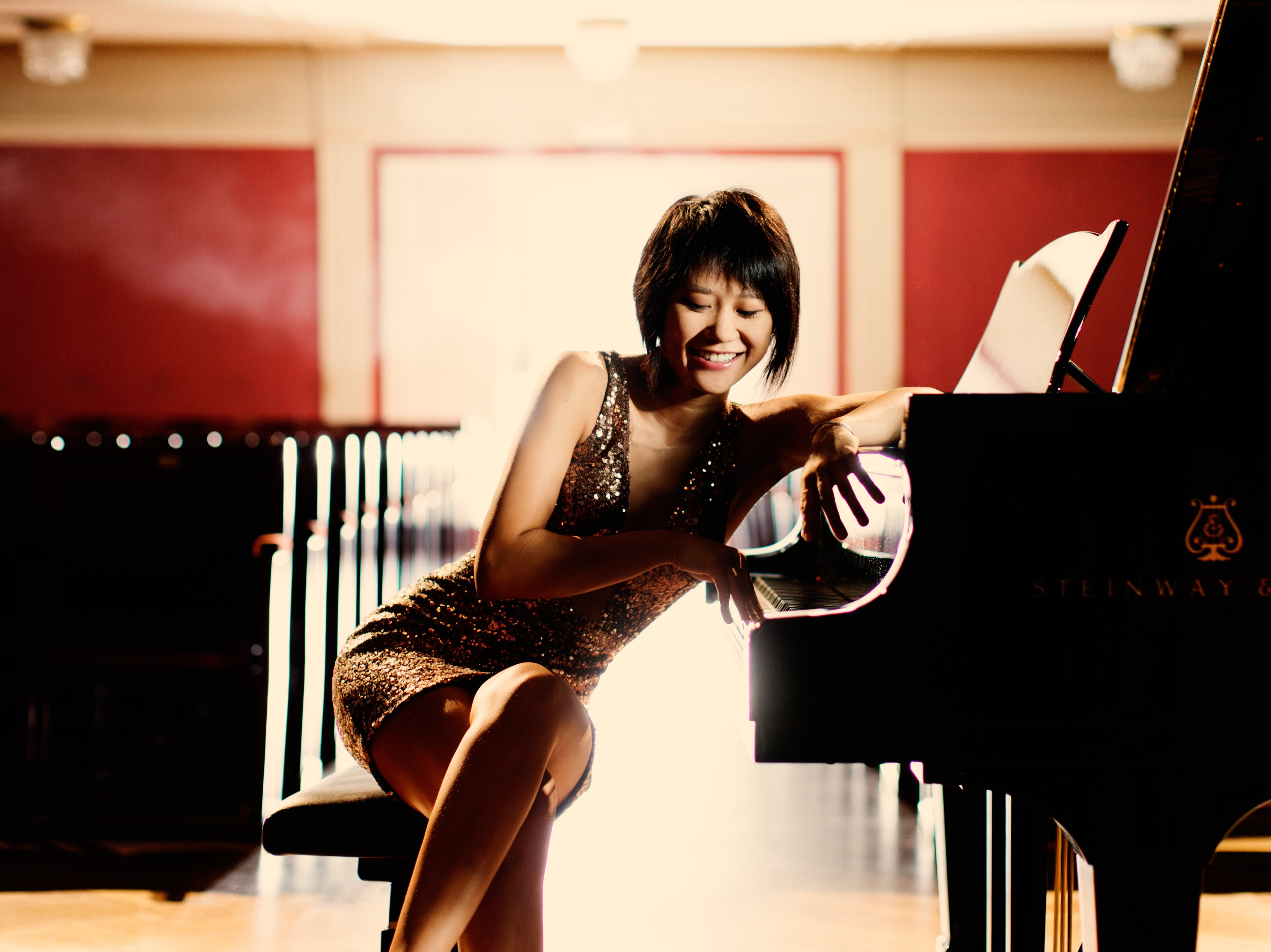 Eine Frau in einem glitzernden Kleid sitzt lächelnd an einem Flügel auf einer schwach beleuchteten Bühne in einem leeren Konzertsaal, wobei warmes Licht sie und den Flügel beleuchtet.