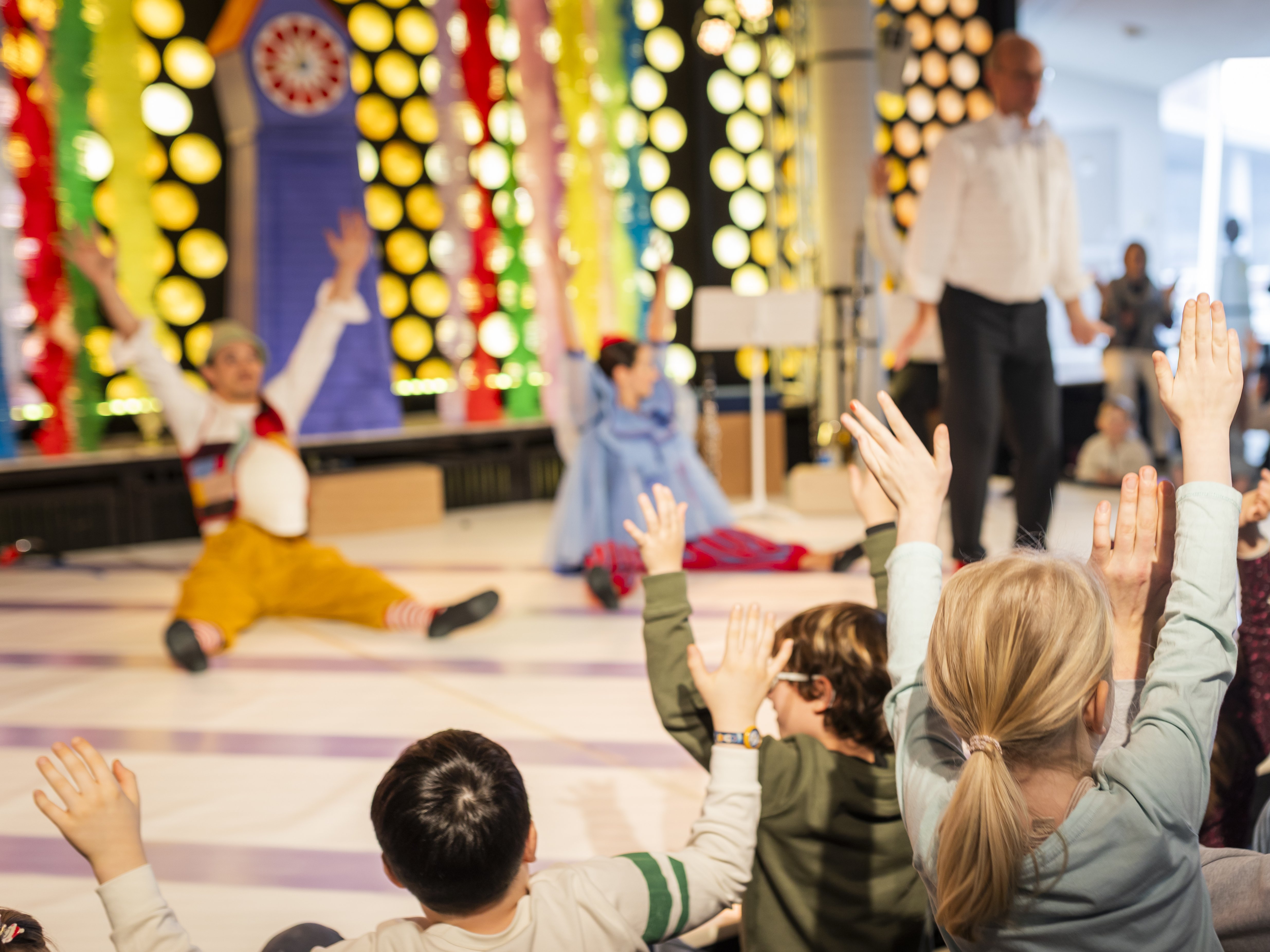 Eine Gruppe von Kindern hebt die Hände, während sie zwei Darsteller auf einer bunt geschmückten Bühne beobachten, während ein Erwachsener sie animiert.