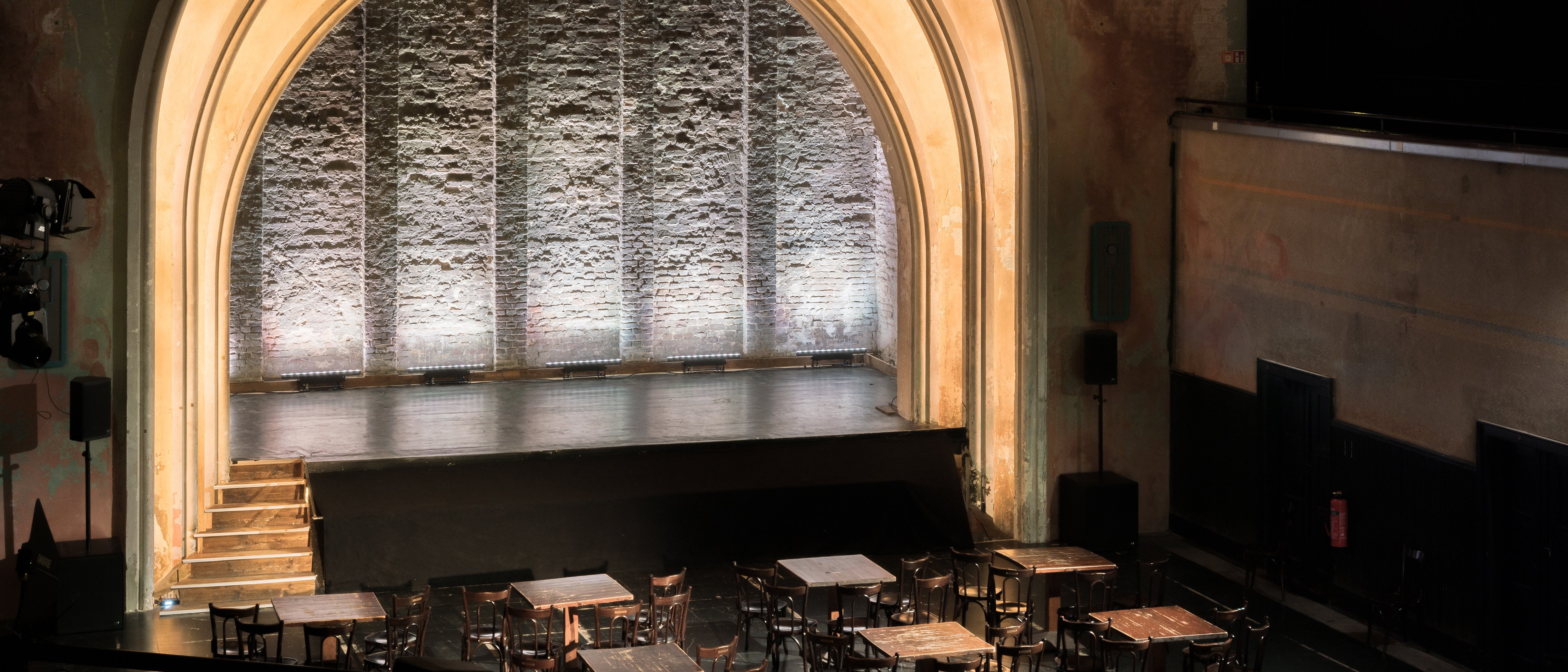 A small, historic event space with an arched stage portal, subtle lighting, and tables with chairs in the audience area.