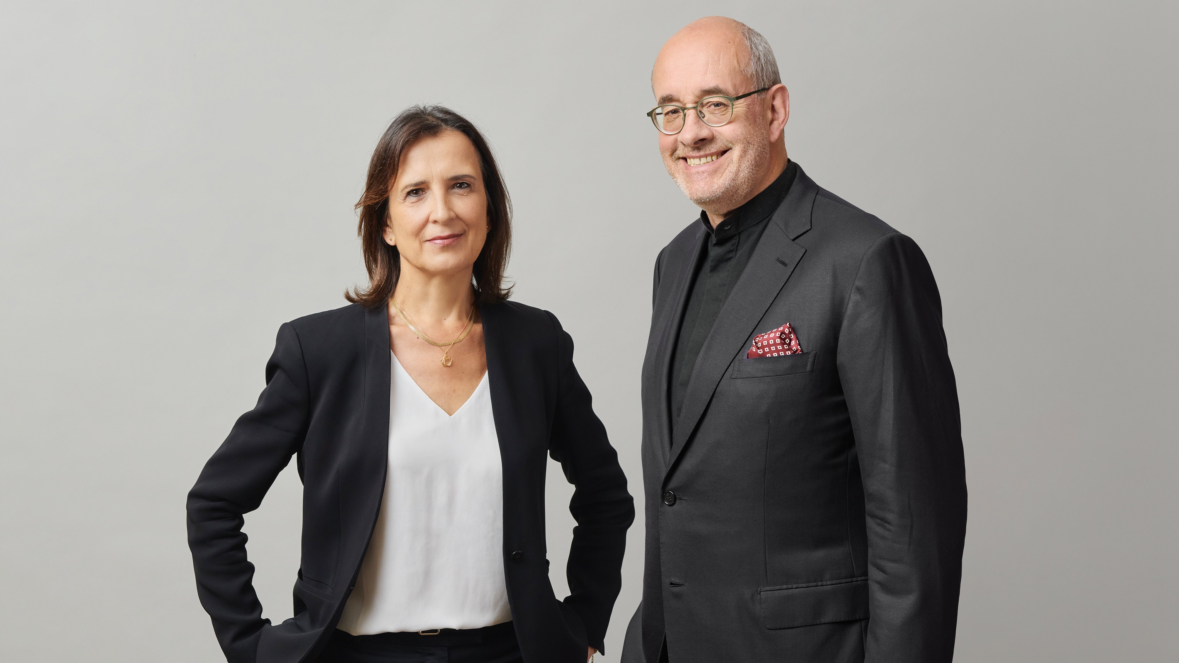 Eine Frau und ein Mann in Geschäftskleidung stehen nebeneinander vor einem schlichten Hintergrund und lächeln beide. Die Frau trägt einen schwarzen Blazer und eine weiße Bluse; der Mann trägt einen schwarzen Anzug mit einem roten Einstecktuch.