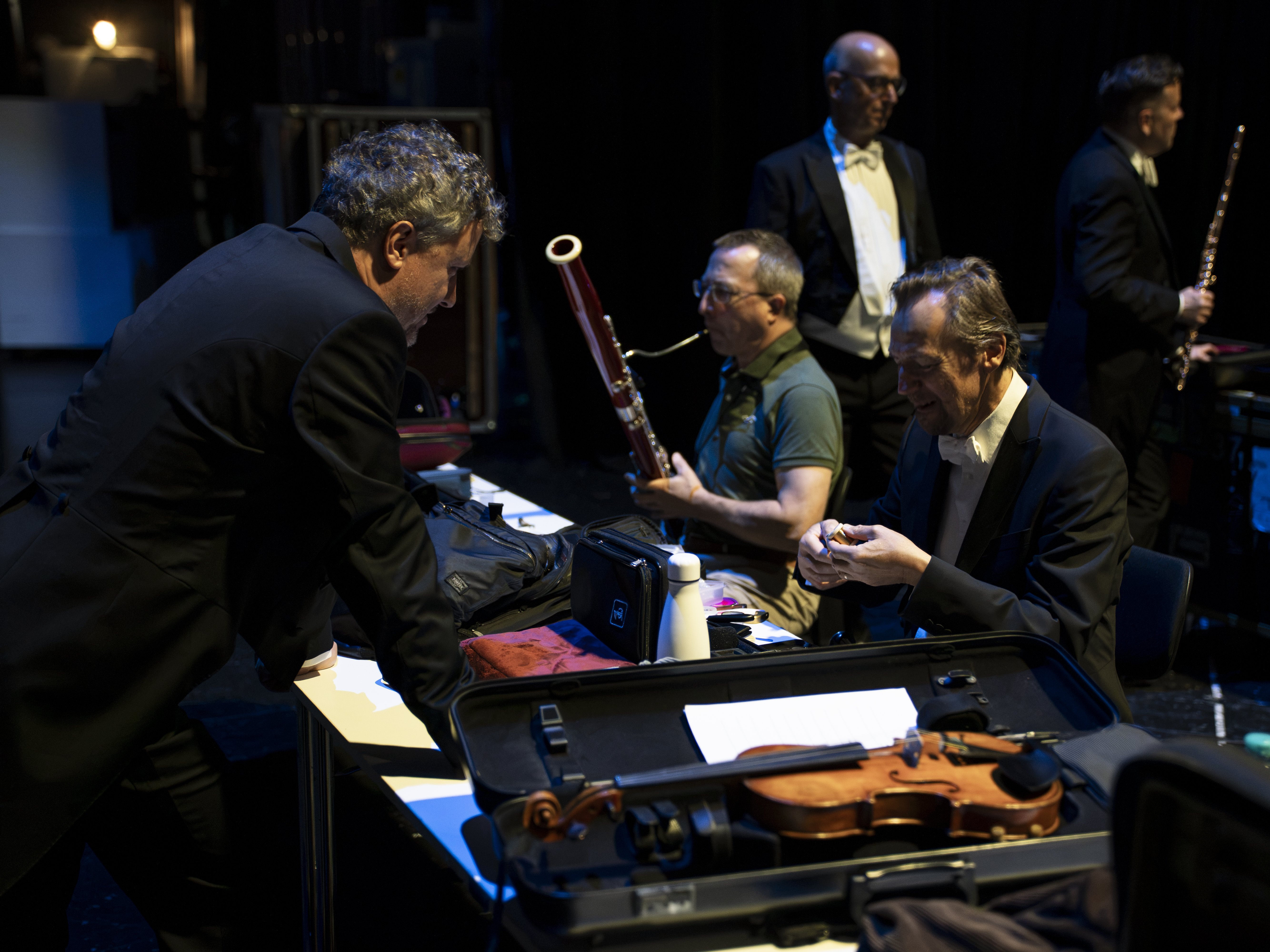 Musicians backstage prepare for a classical concert, handling instruments like a violin and bassoon while dressed formally.