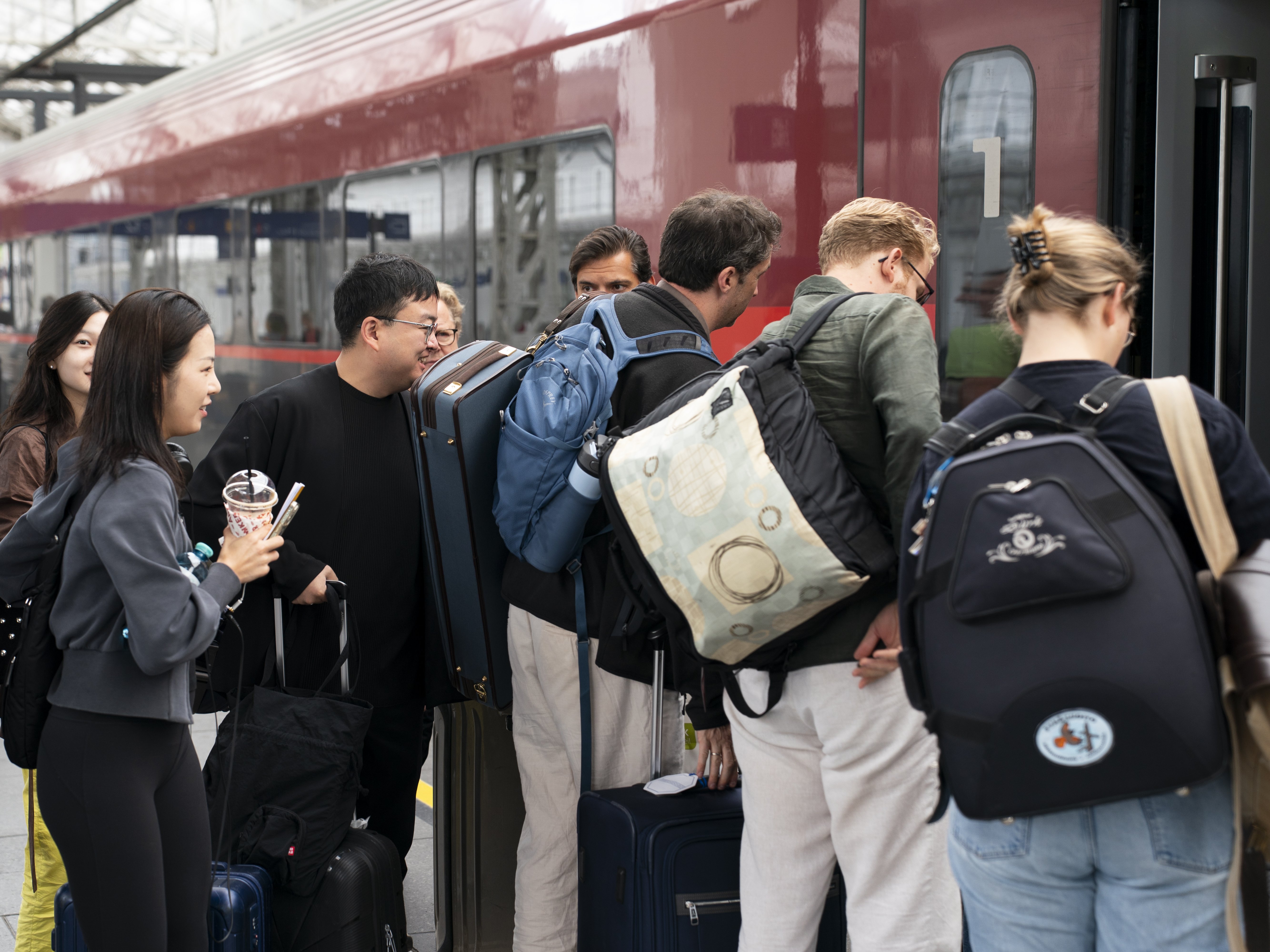 People with backpacks and suitcases board a red train at a station. Some stand in line, while others converse. The platform is busy, the atmosphere lively, and the train door is open.