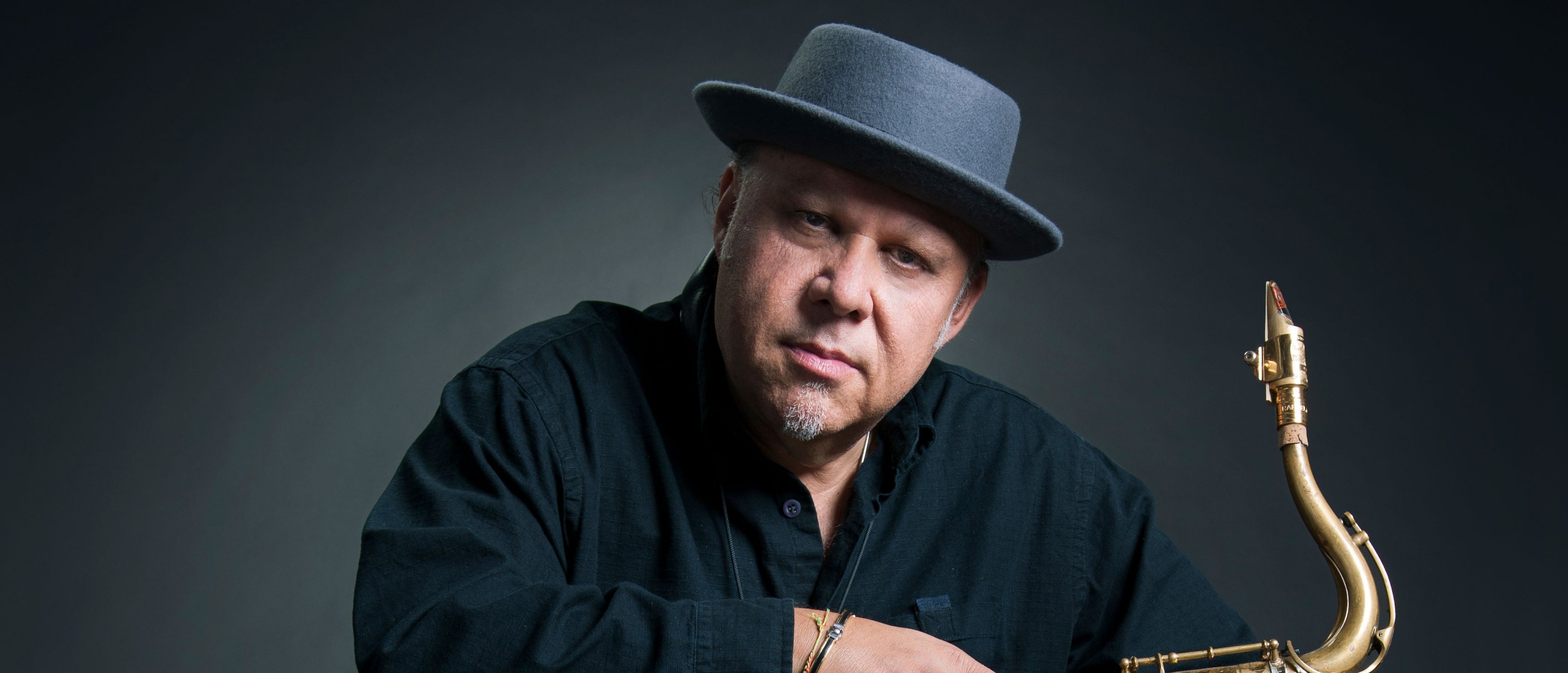 Ein Mann in dunkler Kleidung und mit einem grauen Hut sitzt auf einem schwarzen Stuhl und hält ein Saxophon auf seinem Schoß vor einem dunklen Hintergrund.
