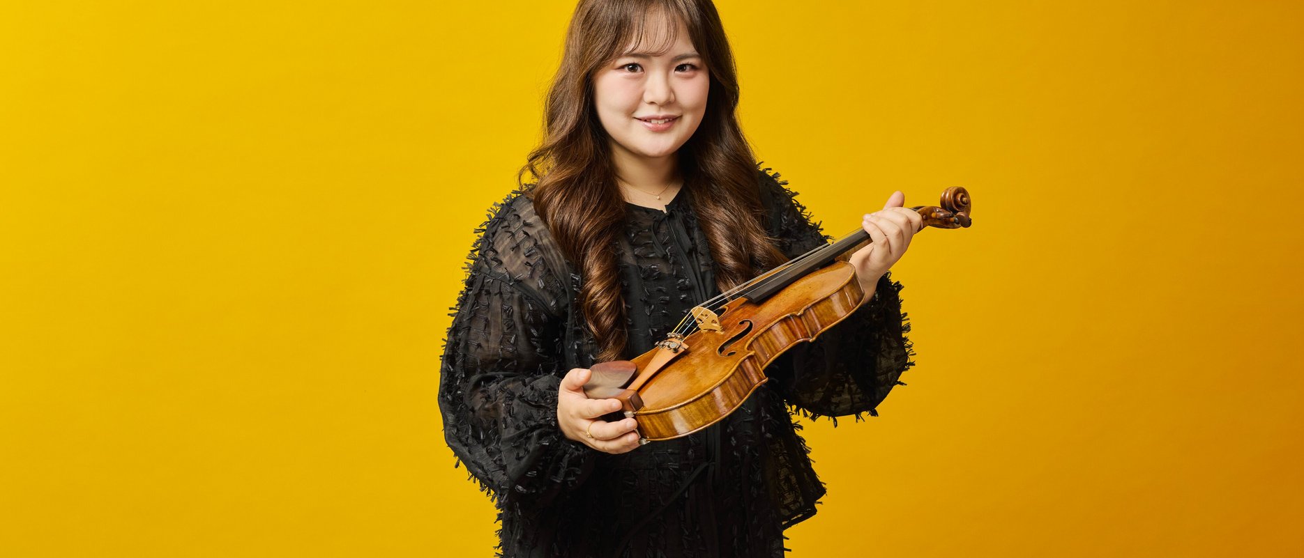 A woman with long brown hair wearing a black top holds a violin and bow, standing and smiling in front of a solid yellow background.