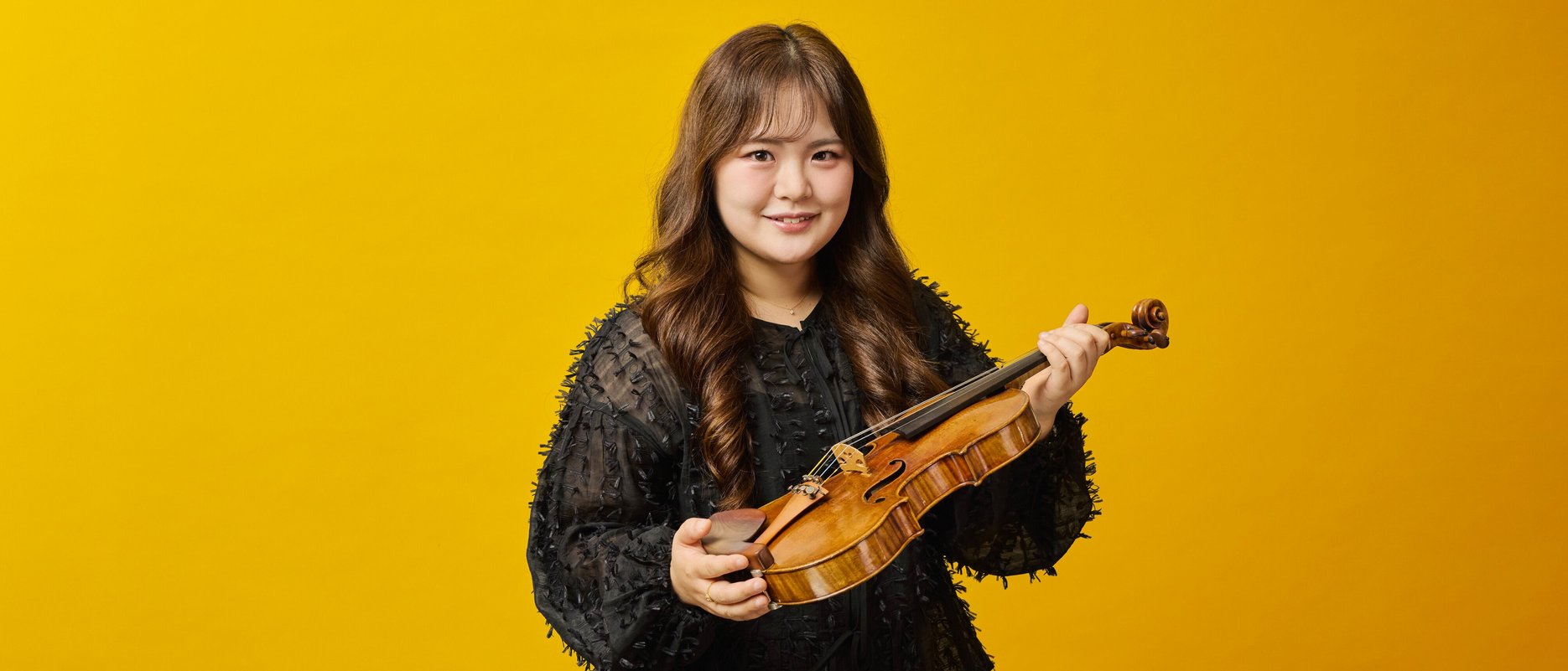 Eine Frau mit langen braunen Haaren, die eine schwarze strukturierte Bluse und eine schwarze Hose trägt, hält eine Geige mit Bogen und lächelt vor einem leuchtend gelben Hintergrund.
