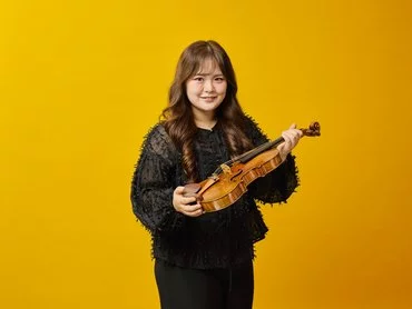 A woman with long brown hair wearing a black top holds a violin and bow, standing and smiling in front of a solid yellow background.