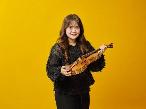 A woman with long brown hair wearing a black top holds a violin and bow, standing and smiling in front of a solid yellow background.