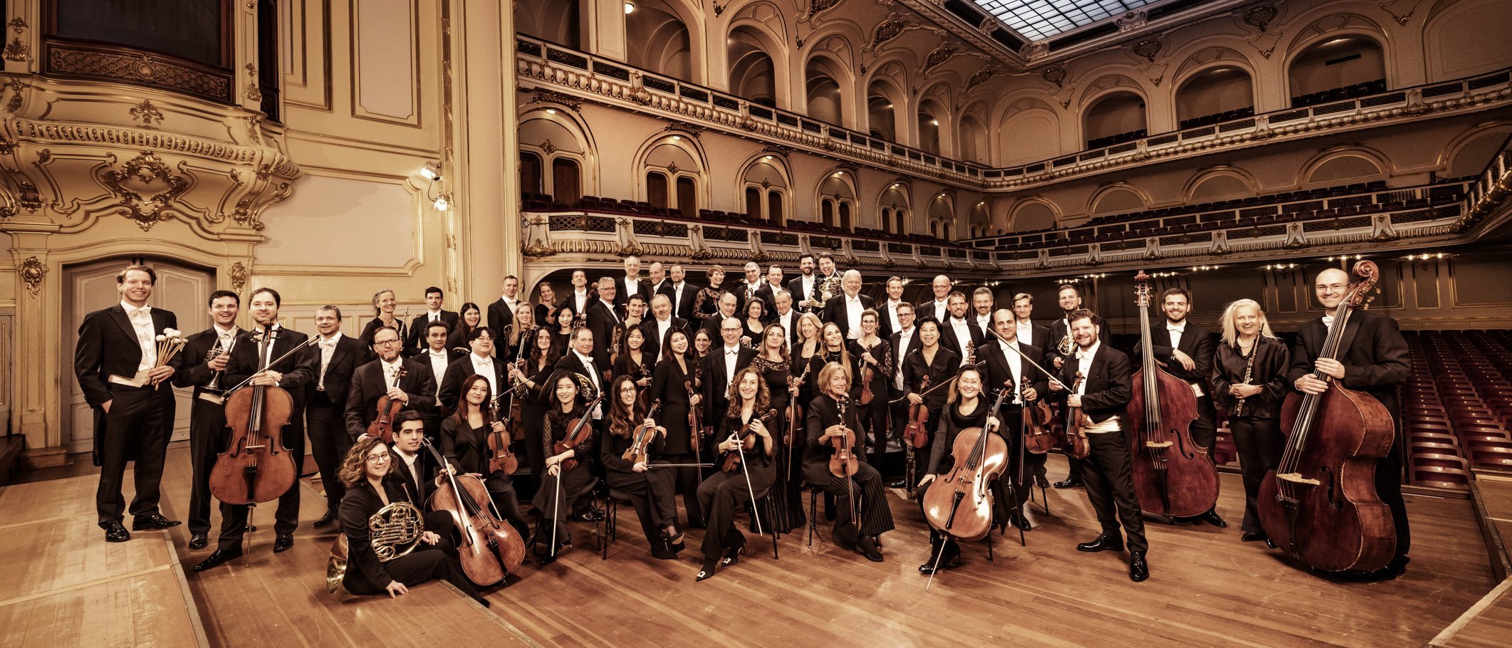 Ein großes Sinfonieorchester in formeller schwarzer Kleidung posiert mit seinen Instrumenten auf der Bühne eines prunkvollen Konzertsaals mit hohen Bögen und einer Glasdecke.