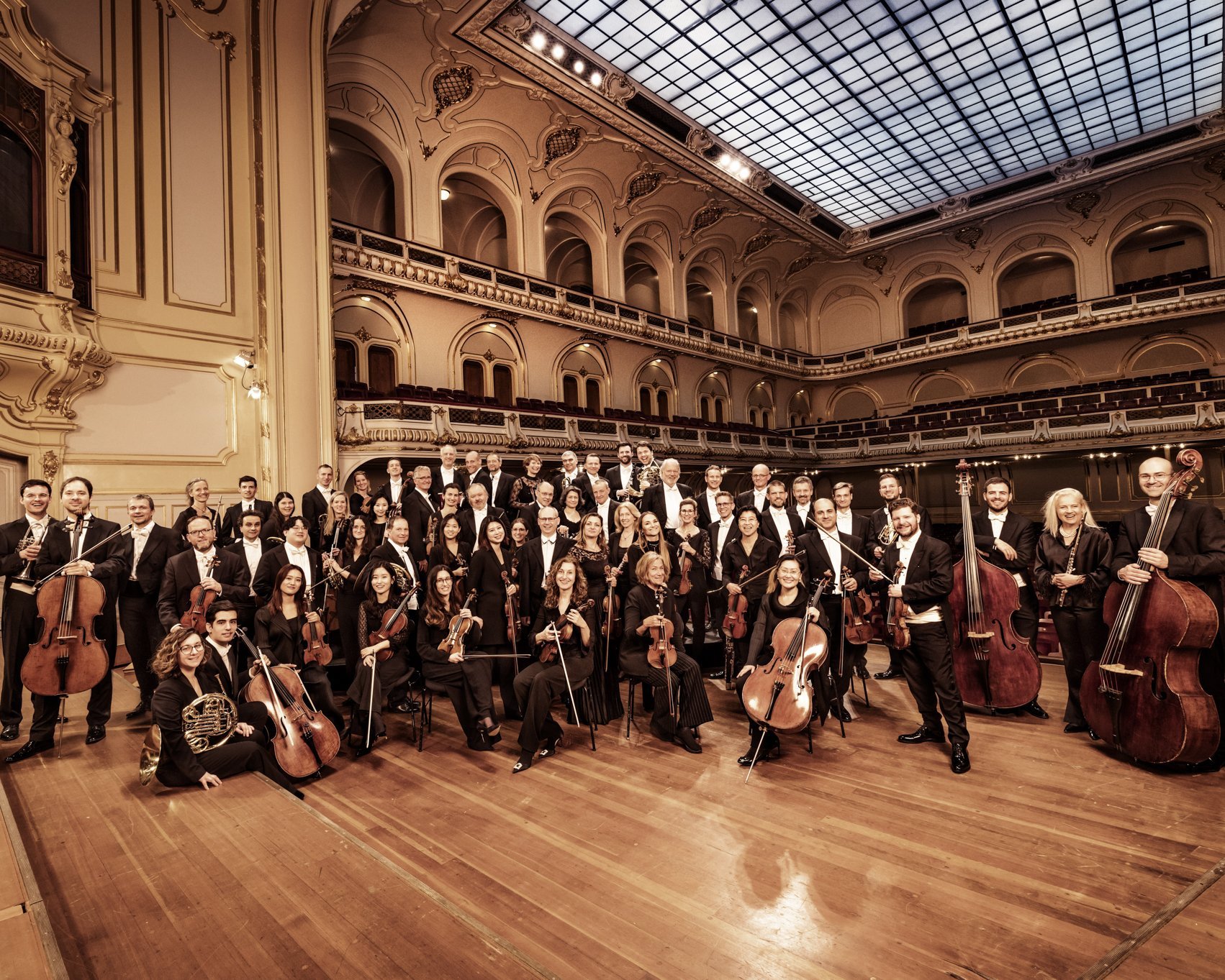 Ein großes Sinfonieorchester in formeller schwarzer Kleidung posiert mit seinen Instrumenten auf der Bühne eines prunkvollen Konzertsaals mit hohen Bögen und einer Glasdecke.