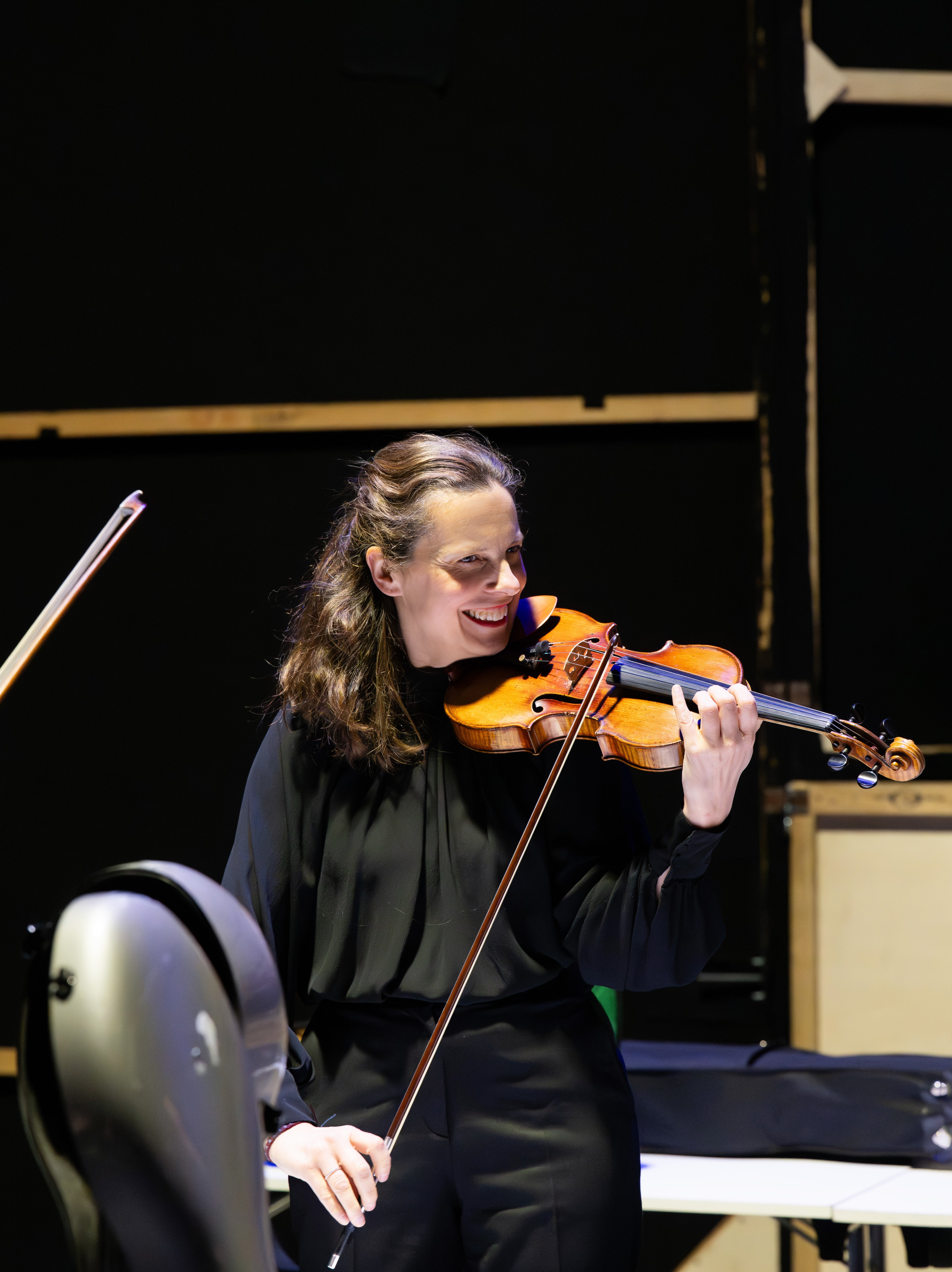 Eine lächelnde, schwarz gekleidete Frau spielt auf der Bühne Geige und hält das Instrument auf der Schulter, während im Vordergrund ein Cello und ein Notenständer teilweise zu sehen sind.