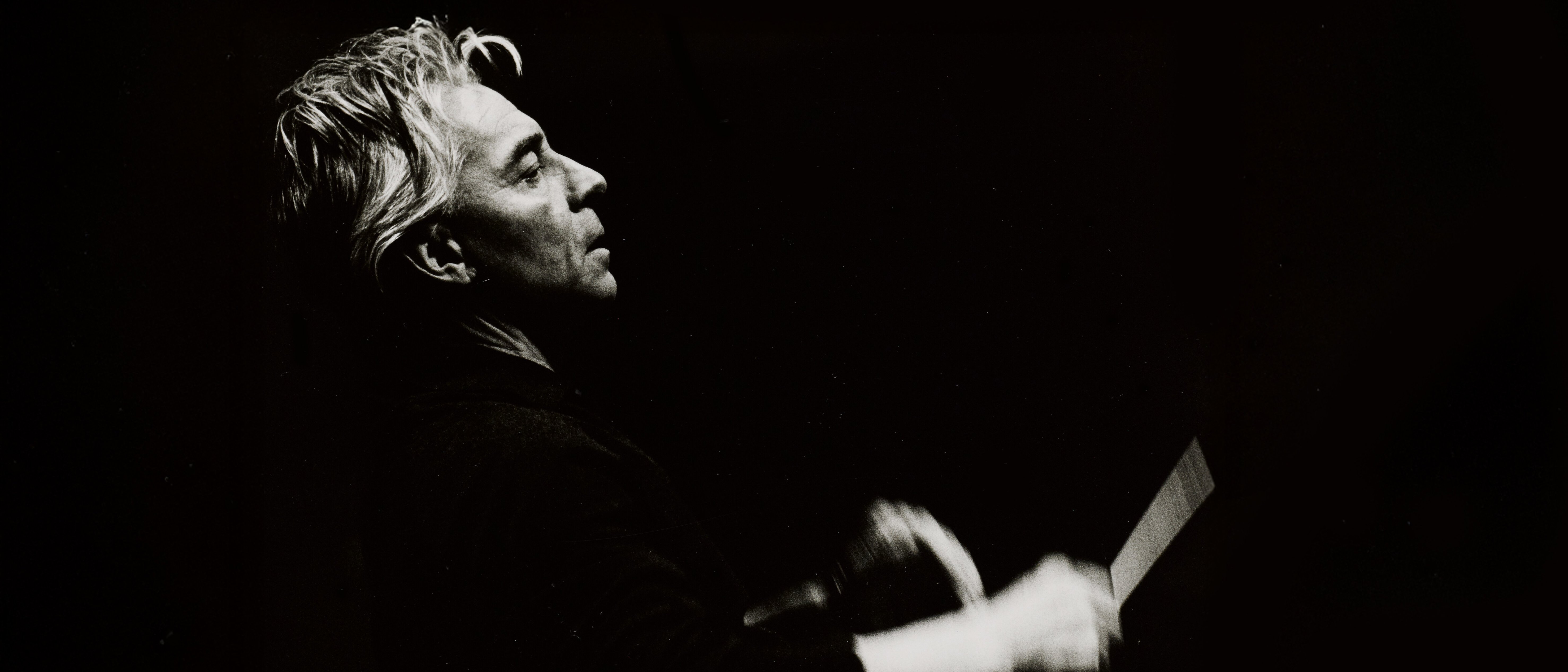 Graustufenfoto eines Mannes mit hellem Haar, im Profil gesehen, der mit einem Taktstock leidenschaftlich Musik dirigiert, vor einem dunklen Hintergrund.