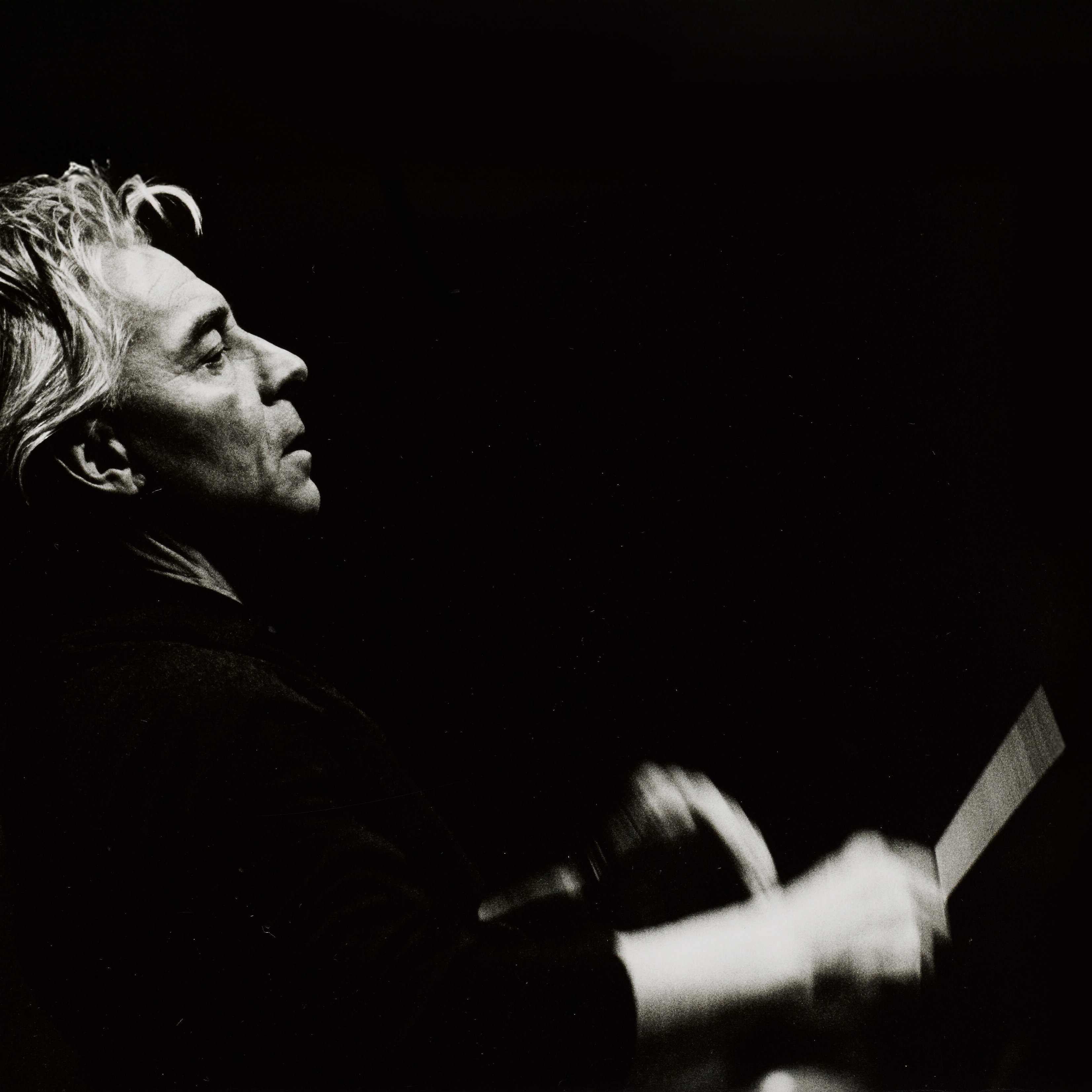 Graustufenfoto eines Mannes mit hellem Haar, im Profil gesehen, der mit einem Taktstock leidenschaftlich Musik dirigiert, vor einem dunklen Hintergrund.