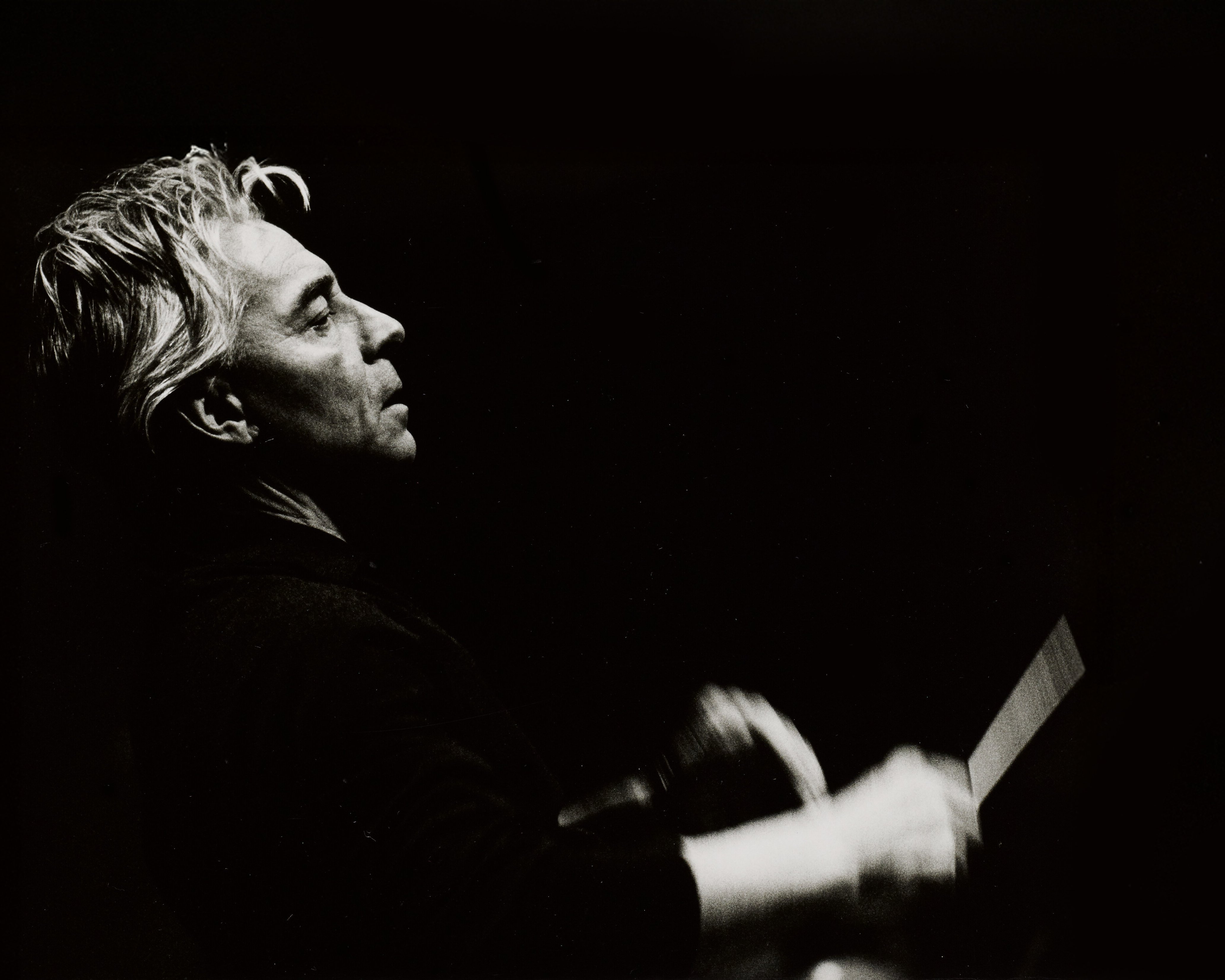 Graustufenfoto eines Mannes mit hellem Haar, im Profil gesehen, der mit einem Taktstock leidenschaftlich Musik dirigiert, vor einem dunklen Hintergrund.