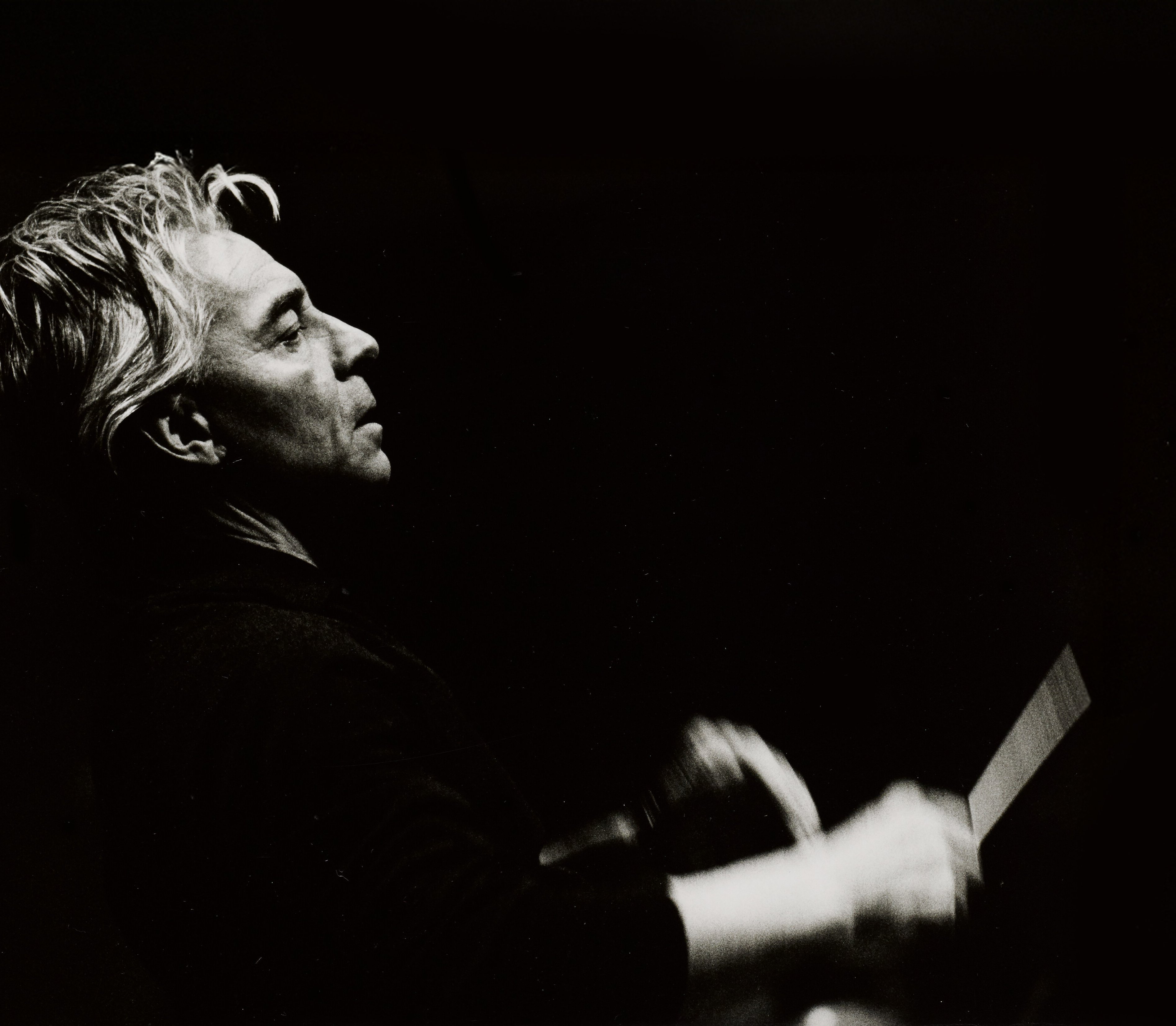 Black-and-white photo of a person with light hair, seen in profile, conducting with an expressive gesture, set against a dark background.