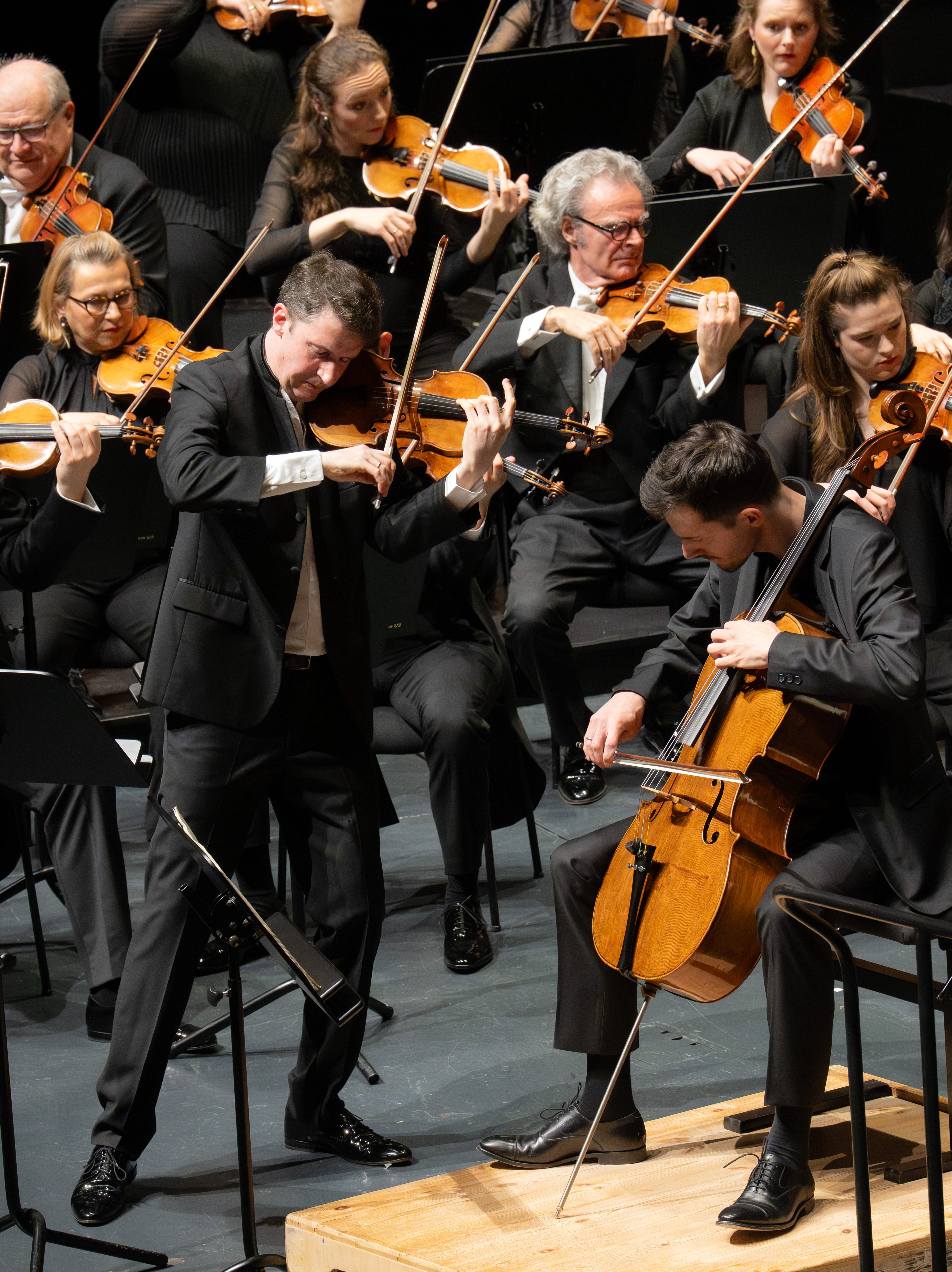 Auf der Bühne spielt ein Geiger gemeinsam mit einem Cellisten solistisch im Vordergrund, während die übrigen Musiker in formeller schwarzer Kleidung sie begleiten.