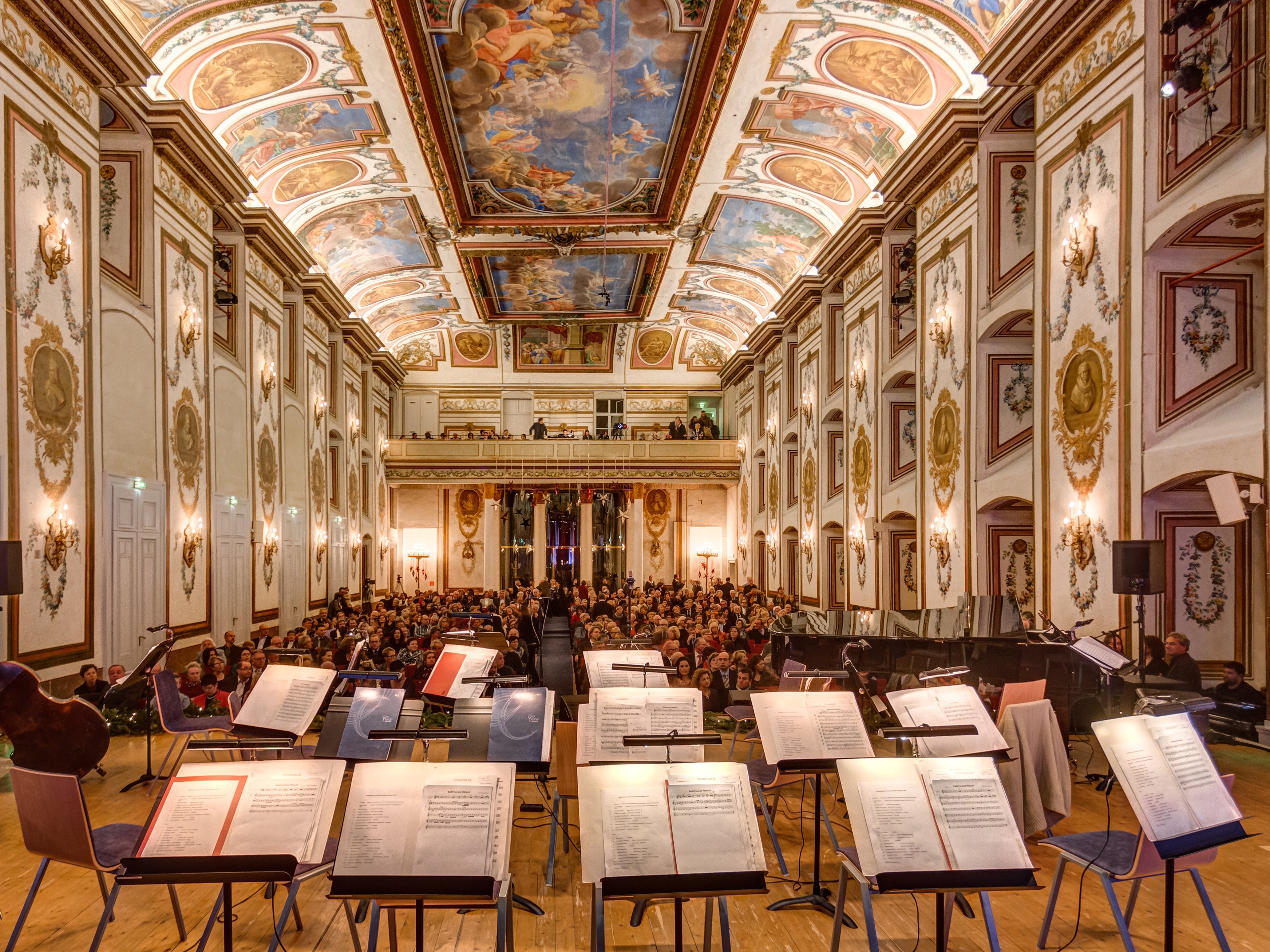 A grand, ornate concert hall with a painted ceiling, golden decorations, and an audience seated before empty music stands on stage, viewed from the performers’ perspective.