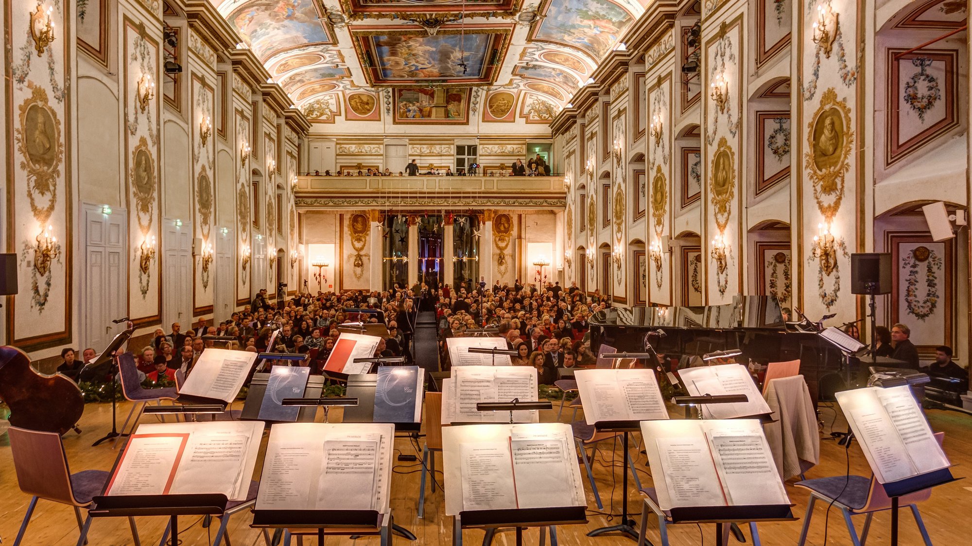 Blick von der Bühne eines großen Konzertsaals mit kunstvoll bemalten Decken, Notenblättern auf Ständern und einem sitzenden Publikum, das auf den Beginn einer Aufführung wartet.