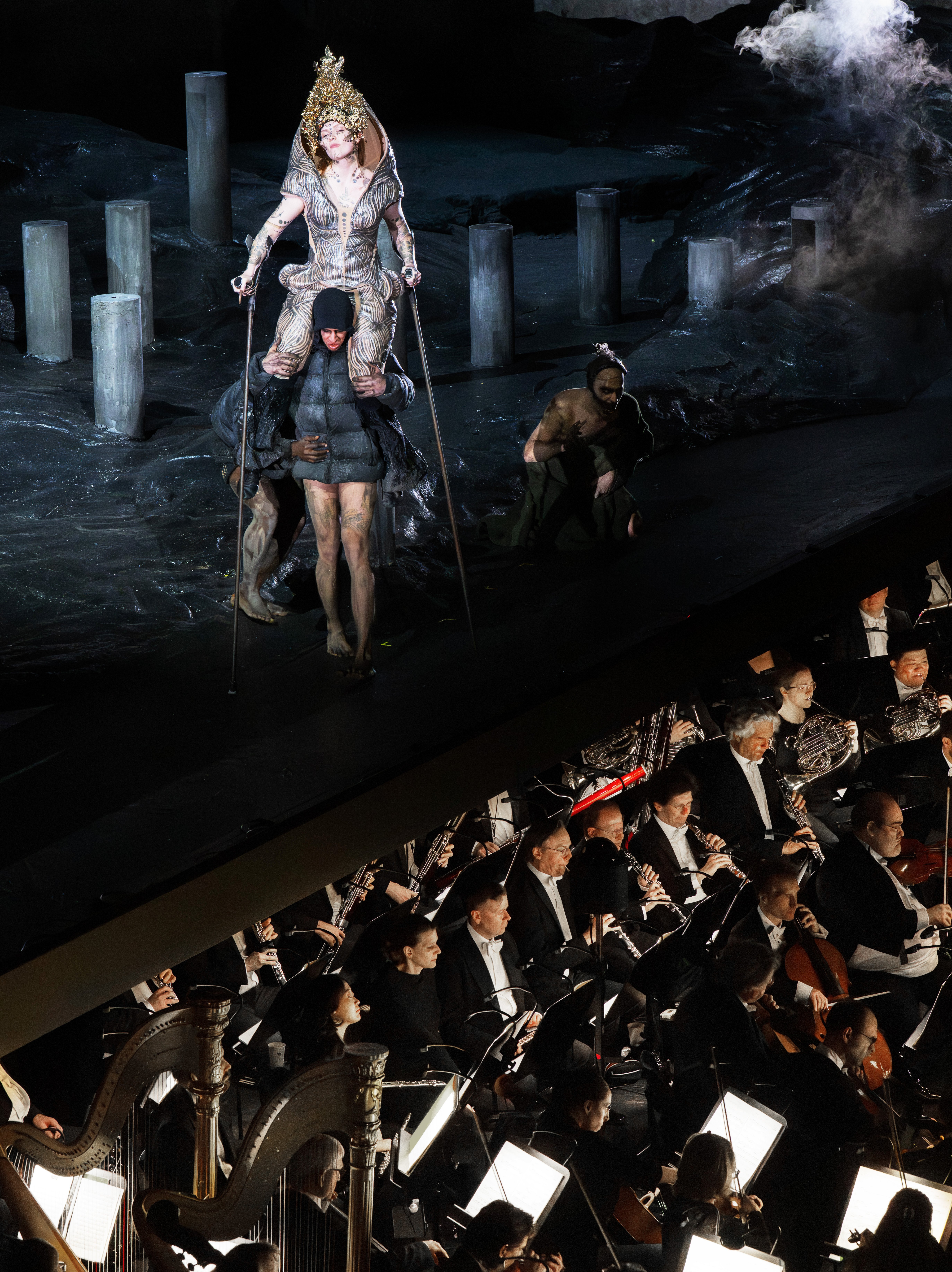 Eine dramatische Bühnenszene mit einem kostümierten Darsteller, der sich über andere erhebt, umgeben von Rauch und Steinsäulen, während unten ein Orchester in formeller Kleidung spielt.