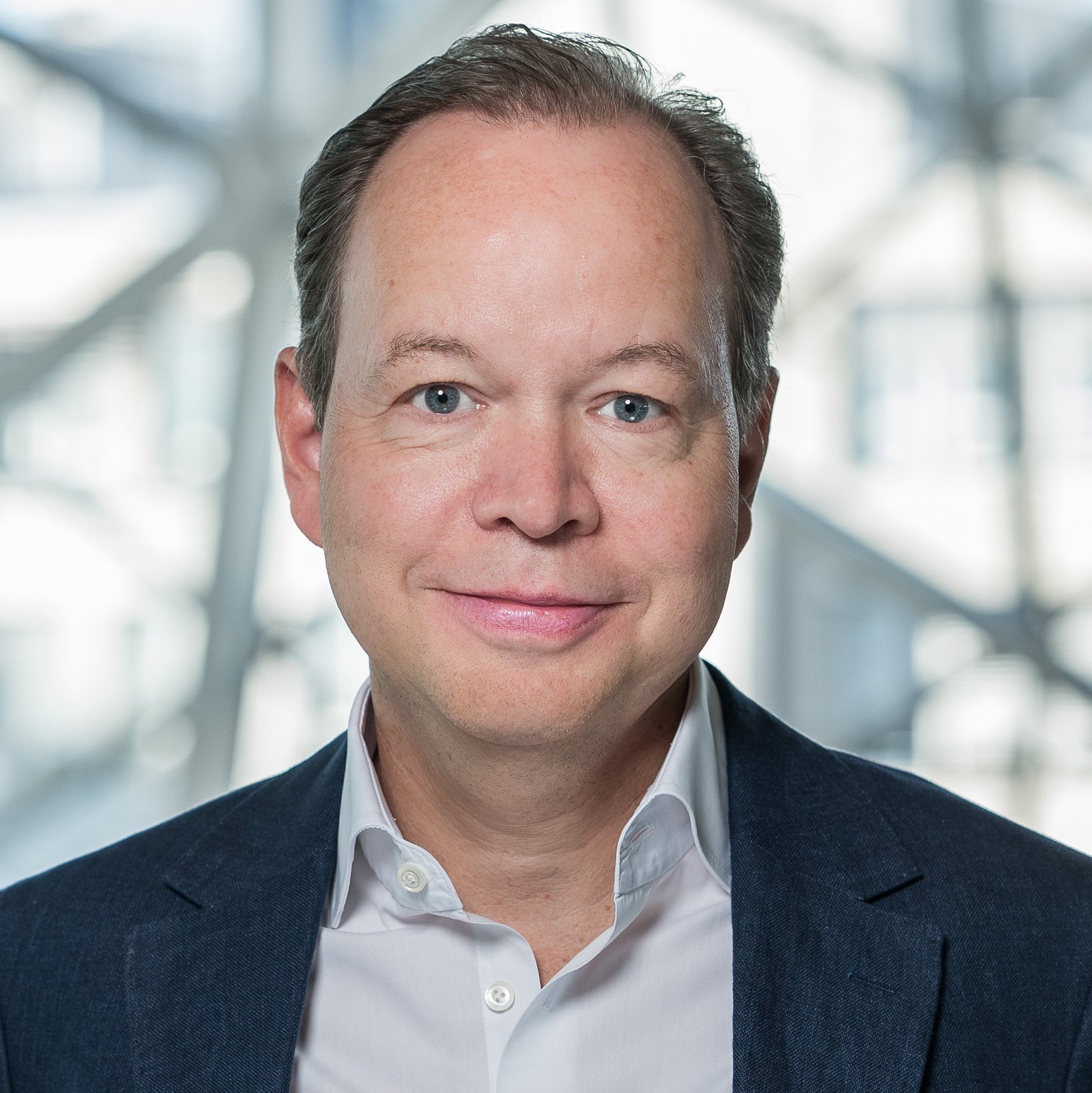 A man in a blue blazer and white shirt stands indoors with a modern glass and steel architectural background, smiling confidently at the camera.
