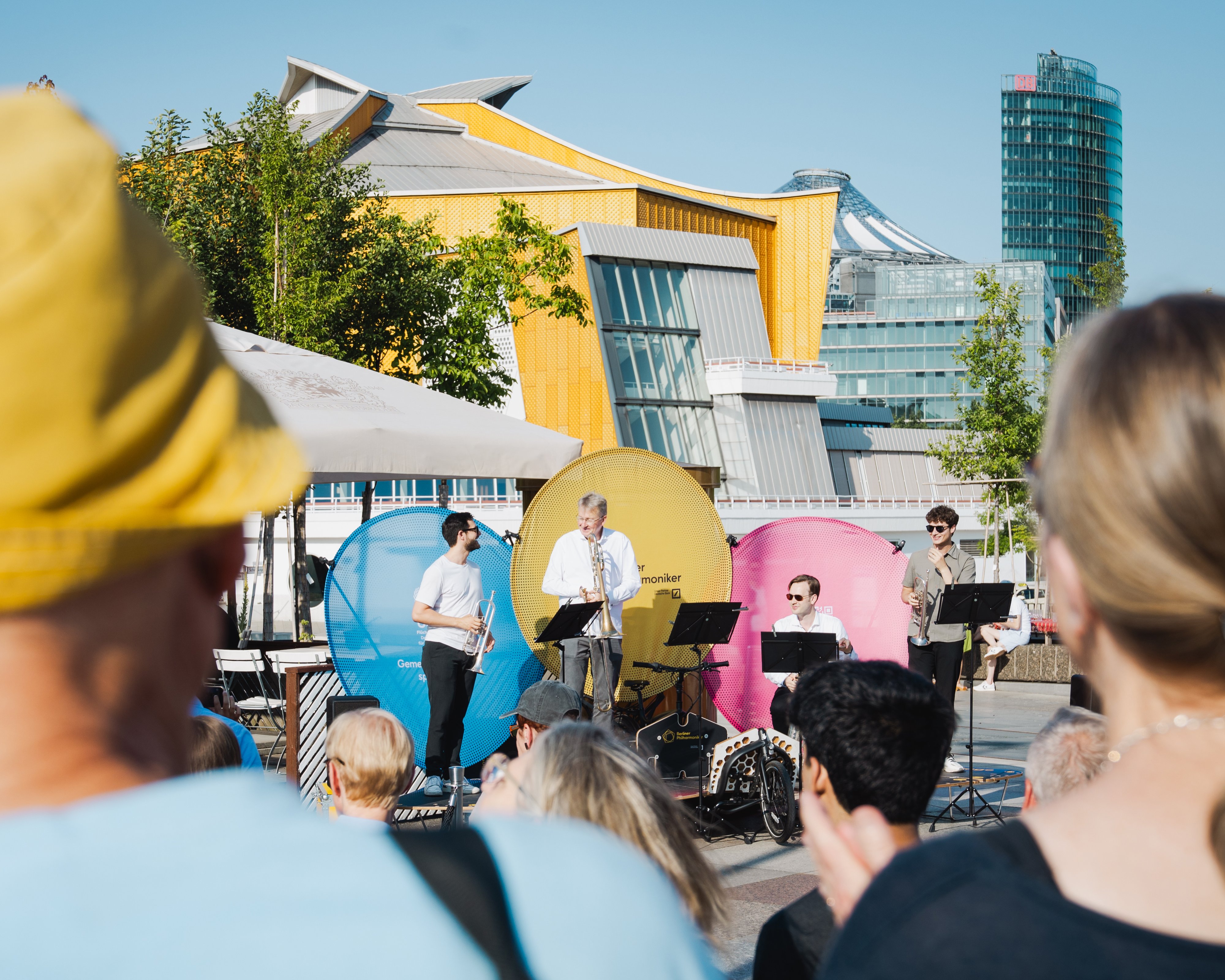 Menschen beobachten ein Konzert auf einer Fahrradbühne im Freien, bei der vier Musiker spielen; die Philharmonie Berlin und Bäume sind unter einem klaren blauen Himmel zu sehen.