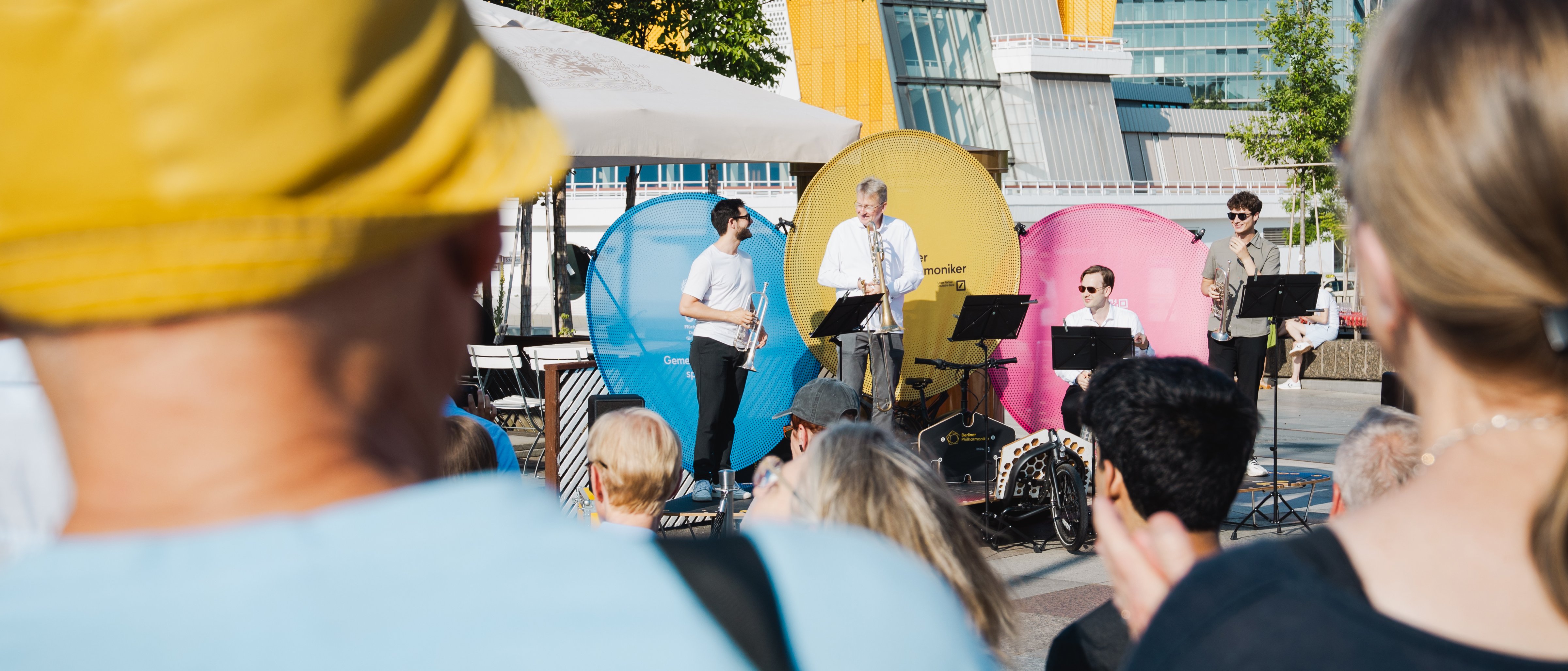 Menschen beobachten ein Konzert auf einer Fahrradbühne im Freien, bei der vier Musiker spielen; die Philharmonie Berlin und Bäume sind unter einem klaren blauen Himmel zu sehen.