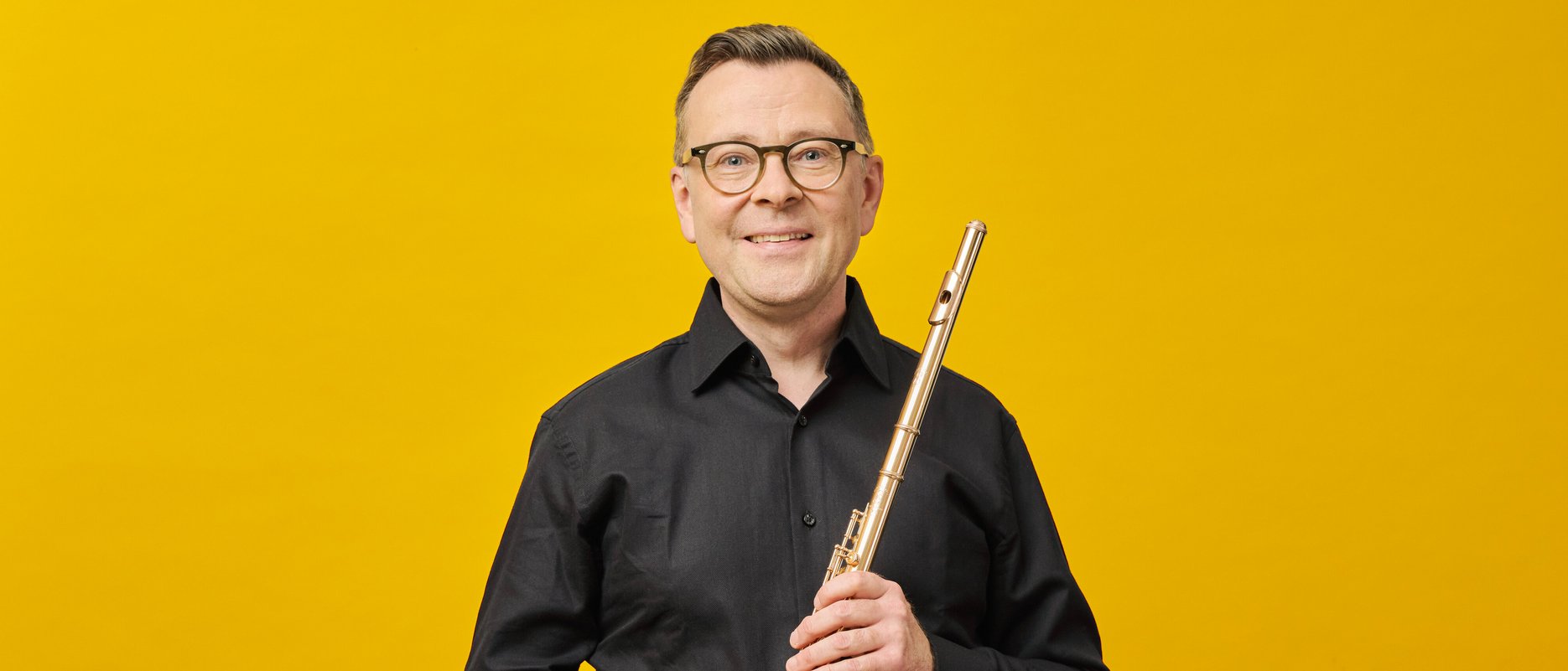 Stefán Ragnar Höskuldsson mit Brille und schwarzem Hemd hält eine Flöte vor einem leuchtend gelben Hintergrund.