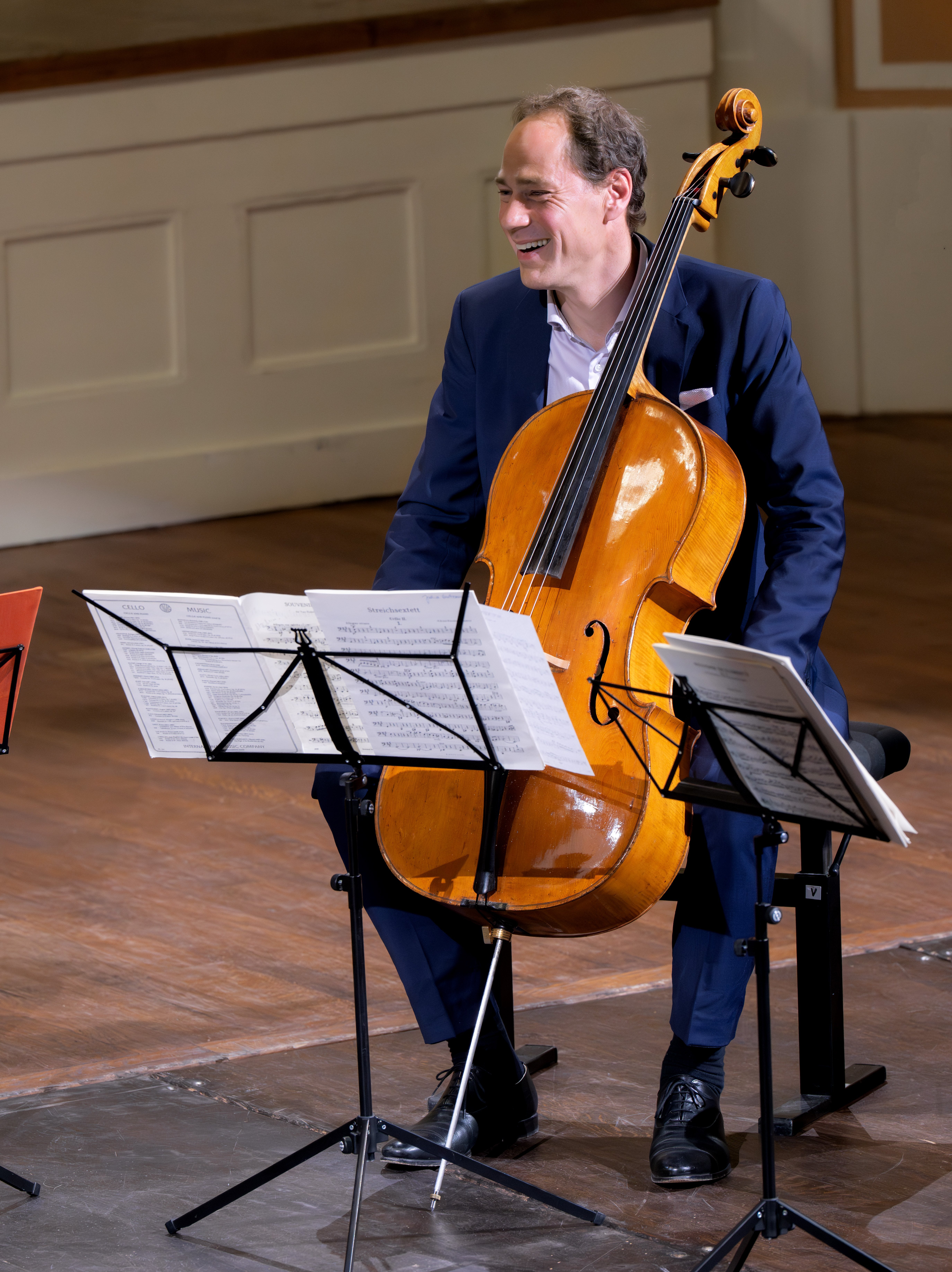 Ein Mann im Anzug sitzt lächelnd mit einem Cello auf der Bühne, umgeben von Notenständern mit Notenblättern, in einem Konzertsaal mit Holzboden und weißen Wänden.