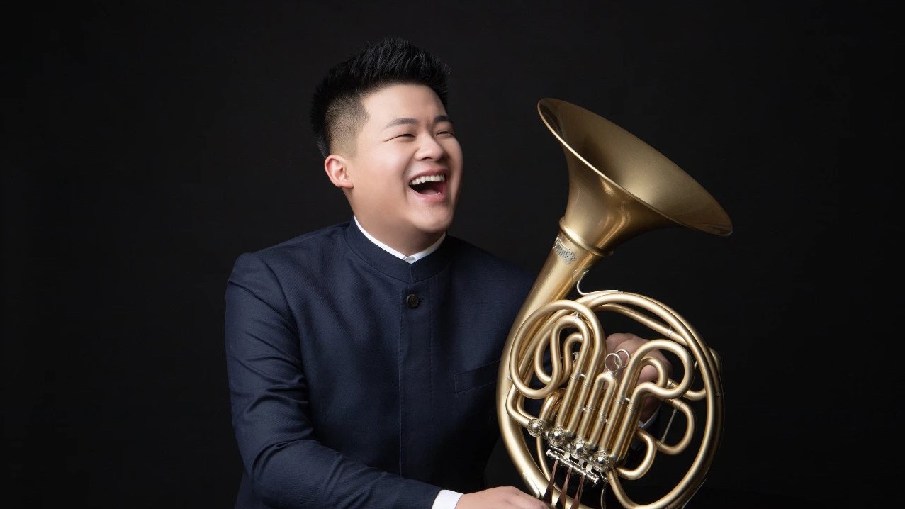 Eine Person in einem dunklen Anzug lacht, während sie ein Waldhorn vor einem schlichten schwarzen Hintergrund hält.