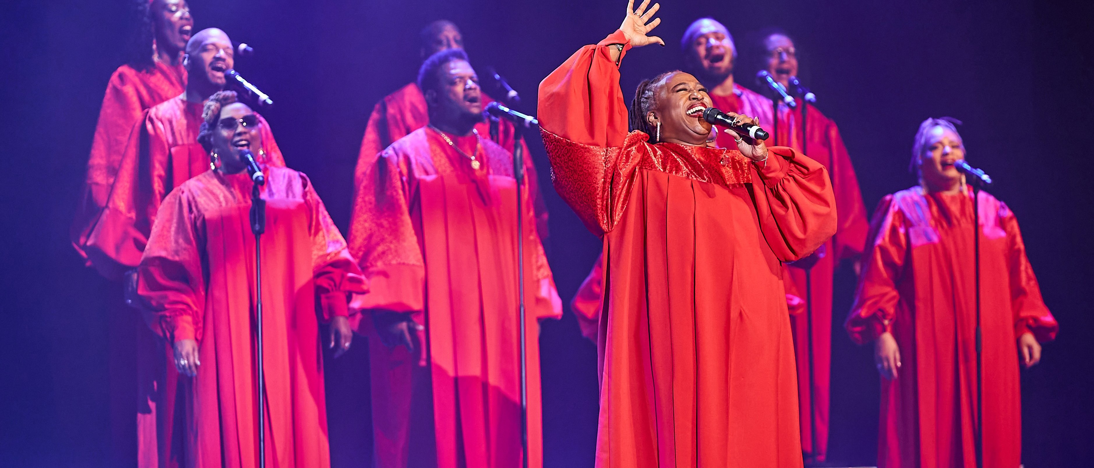 Harlem Gospel Singers in passenden roten Gewändern singt leidenschaftlich auf der Bühne, wobei eine Solistin vorne die Hand hebt, während andere im Hintergrund harmonieren, alles beleuchtet von Bühnenscheinwerfern.