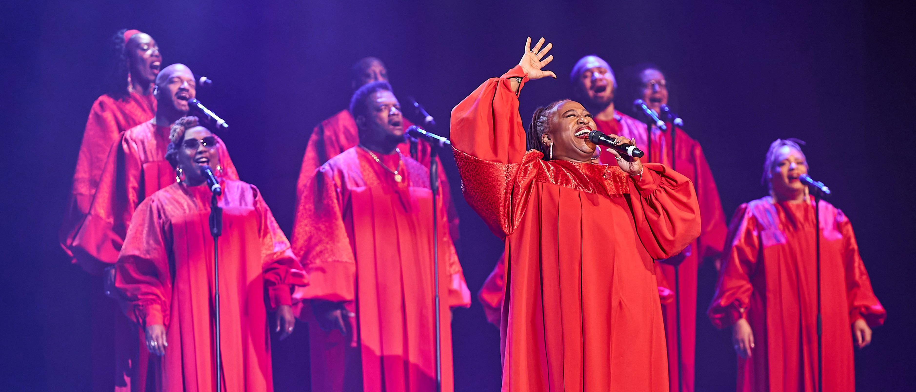 Harlem Gospel Singers in passenden roten Gewändern singt leidenschaftlich auf der Bühne, wobei eine Solistin vorne die Hand hebt, während andere im Hintergrund harmonieren, alles beleuchtet von Bühnenscheinwerfern.