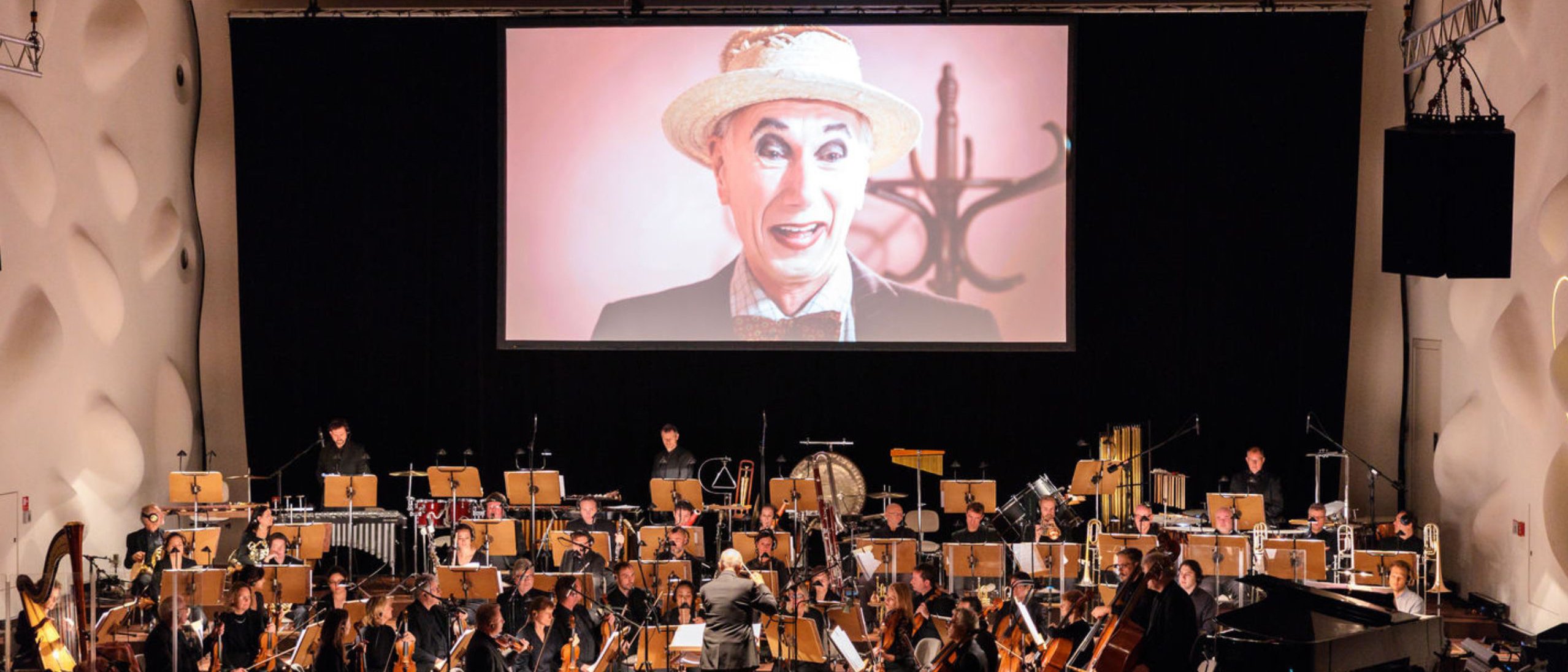 Ein Dirigent leitet ein Orchester auf der Bühne. Hinter ihm ist auf einer großen Leinwand eine Person mit Strohhut und Fliege zu sehen, die mit übertriebenem Make-up lächelt.