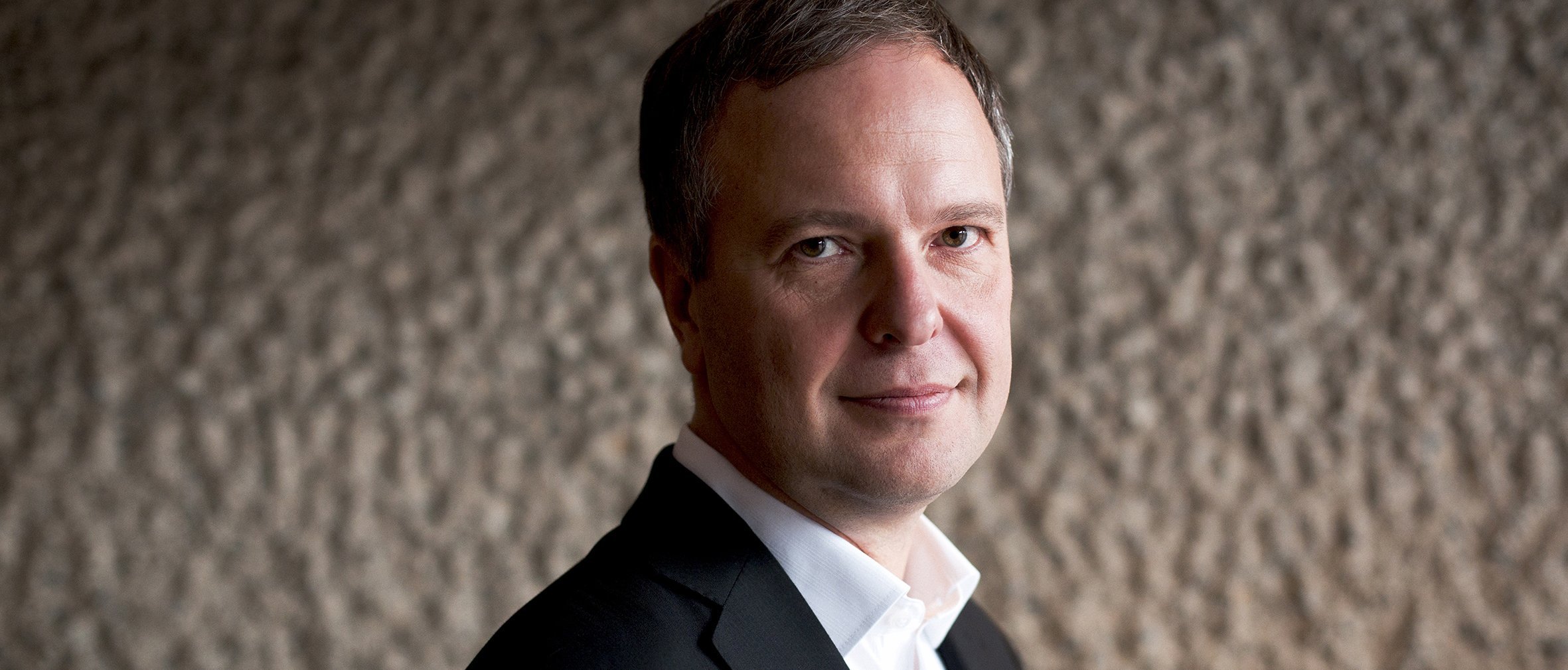 Sakari Oramo in schwarzem Anzug und weißem Hemd steht vor einer strukturierten Wand und blickt mit einem leichten Lächeln in die Kamera. Der Hintergrund weist eine raue, steinartige Oberfläche auf.