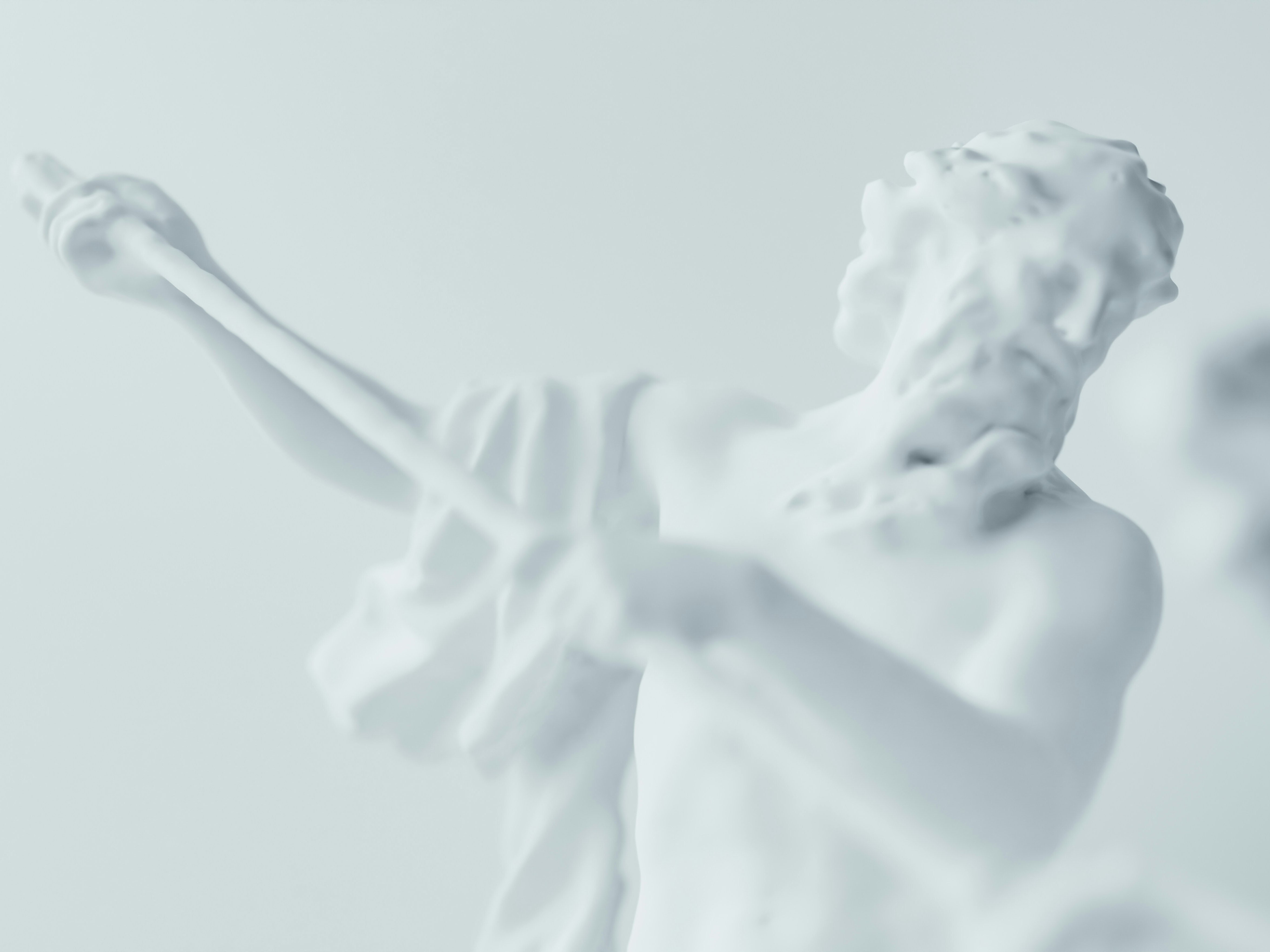 A close-up of a white marble statue depicting a bearded figure holding a staff, with blurred details and another statue partially visible in the foreground, all against a pale background.
