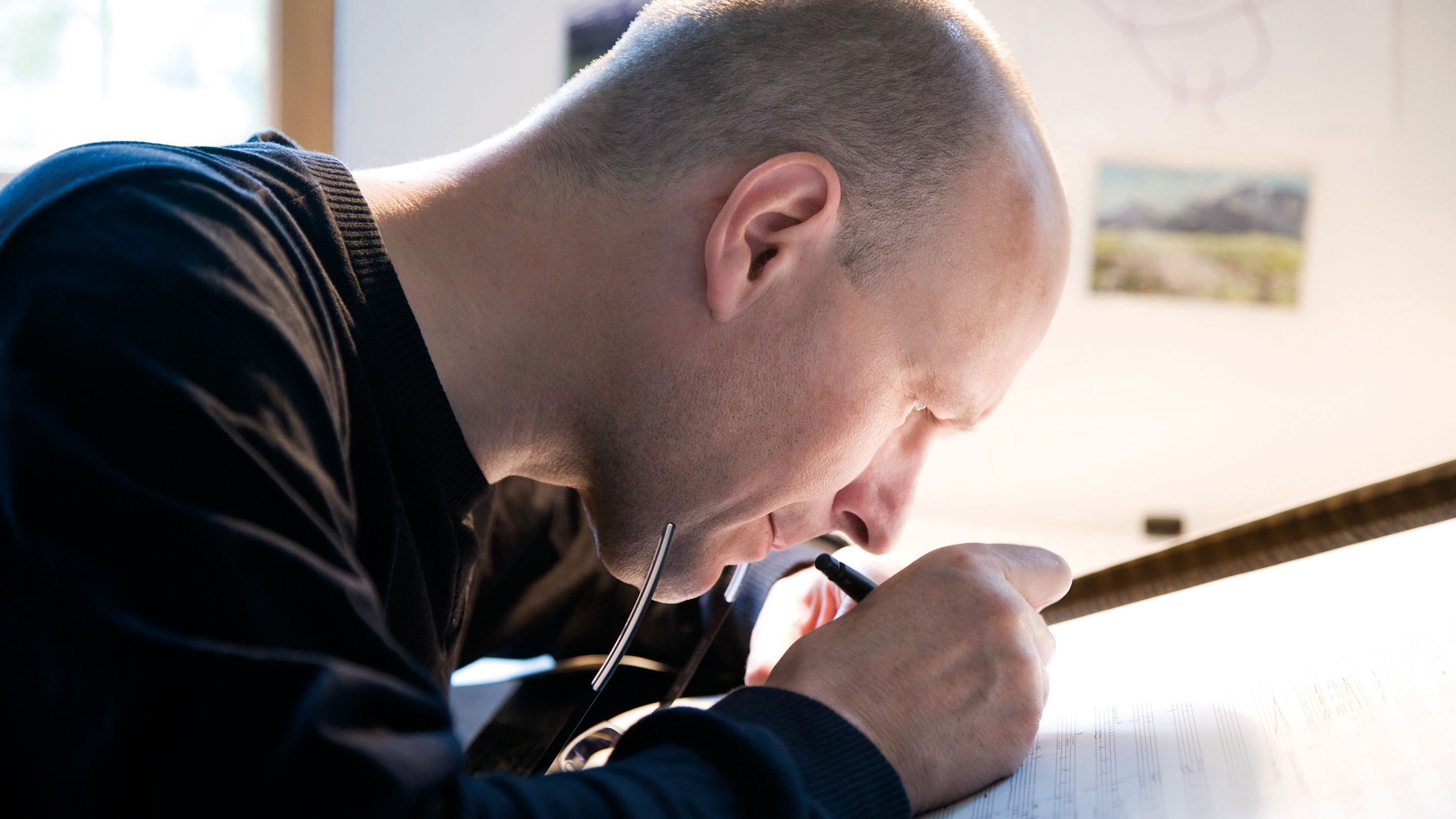 Eine Person mit Glatze sitzt an einem gut beleuchteten Schreibtisch und schreibt oder zeichnet konzentriert auf ein großes Blatt Papier, wobei sie in der einen Hand eine Brille und in der anderen einen Stift hält.