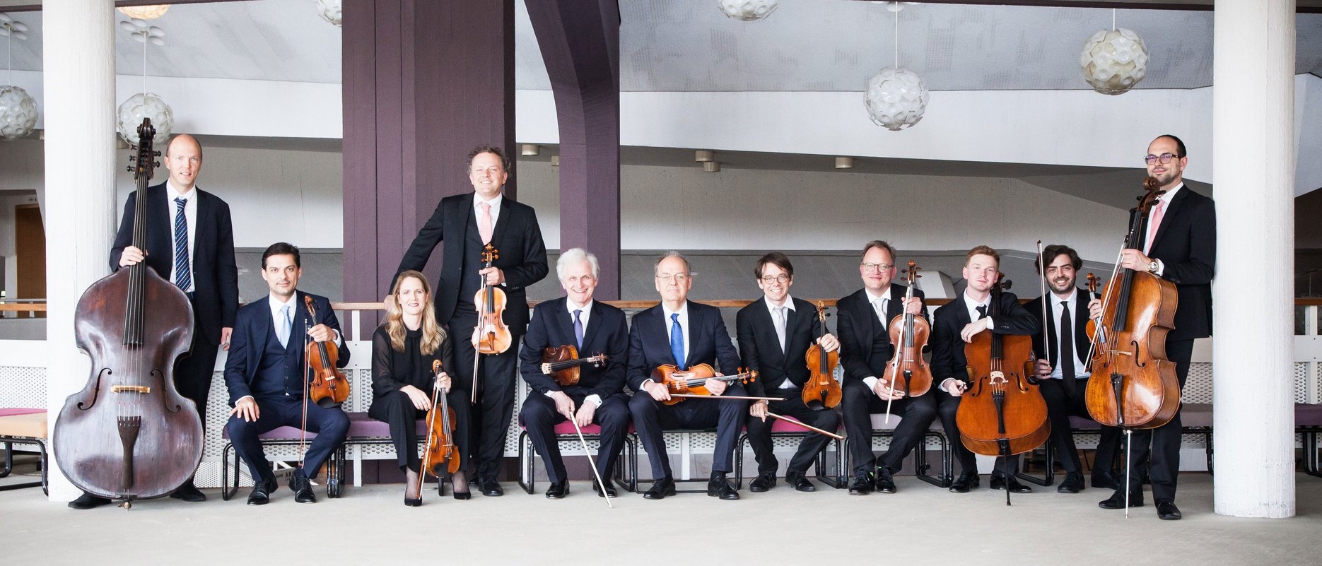 Zehn Musiker in formeller Kleidung posieren mit Streichinstrumenten, darunter Geigen, Bratschen, Celli und ein Kontrabass, in einem modernen Gebäude mit weißen Wänden und hängenden runden Lichtern.