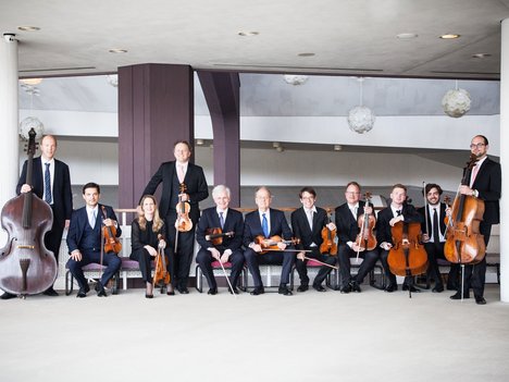 A group of eleven musicians in formal attire pose indoors with string instruments, including violins, violas, cellos, and a double bass, against a modern architectural backdrop.