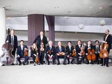 Zehn Musiker in formeller Kleidung posieren mit Streichinstrumenten, darunter Geigen, Bratschen, Celli und ein Kontrabass, in einem modernen Gebäude mit weißen Wänden und hängenden runden Lichtern.