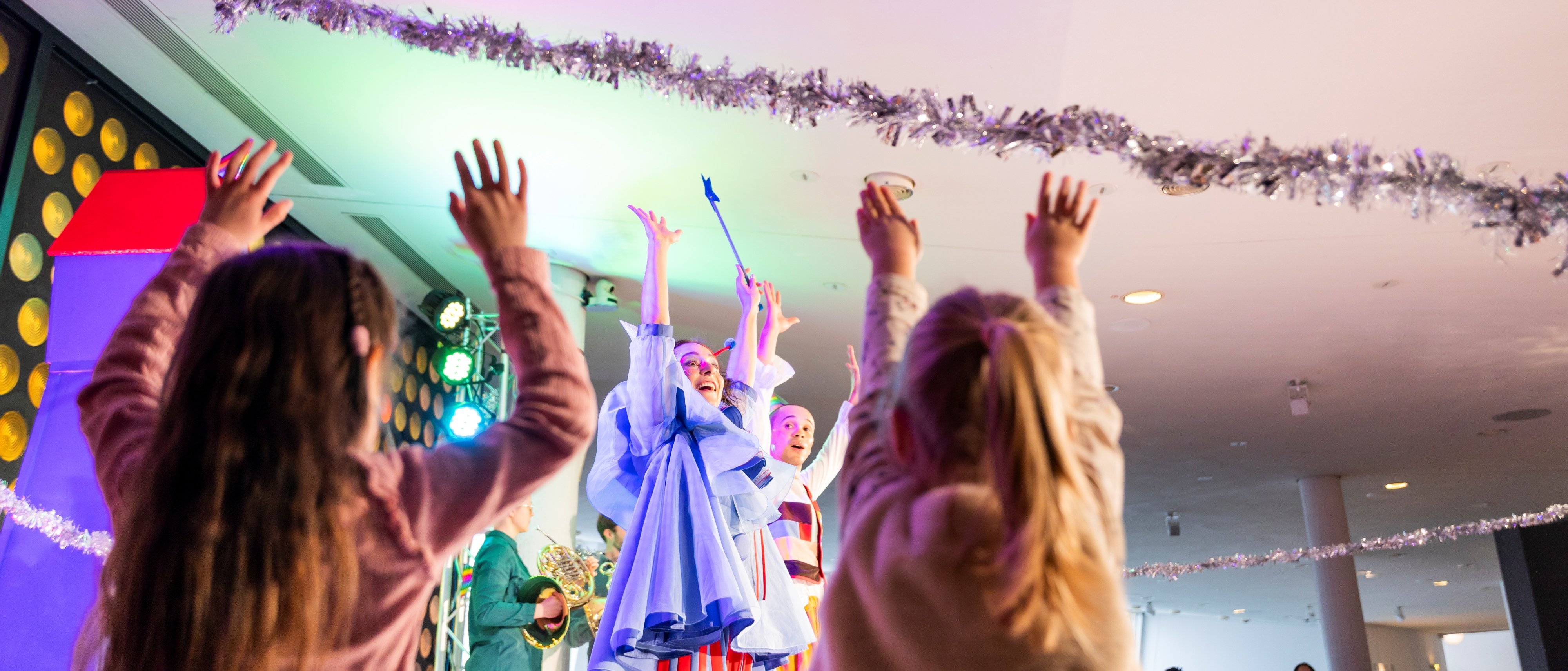 Kinder mit erhobenen Armen beobachten Darsteller, die als farbenfrohe Figuren verkleidet auf einer geschmückten Bühne mit festlichen Girlanden und hellen Lichtern im Hintergrund während einer lebhaften Veranstaltung oder Show auftreten.