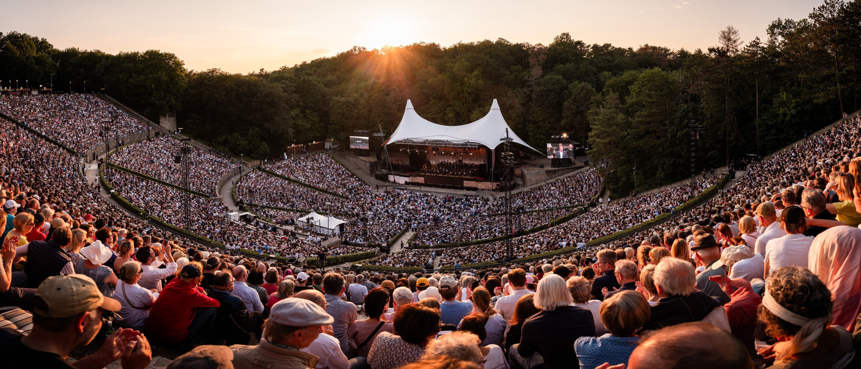 Ein großes Amphitheater unter freiem Himmel, vollgepackt mit Menschen, die ein Konzert bei Sonnenuntergang verfolgen. Die Bühne ist von einem weißen Baldachin bedeckt, umgeben von üppig grünen Bäumen unter einem warmen Himmel.