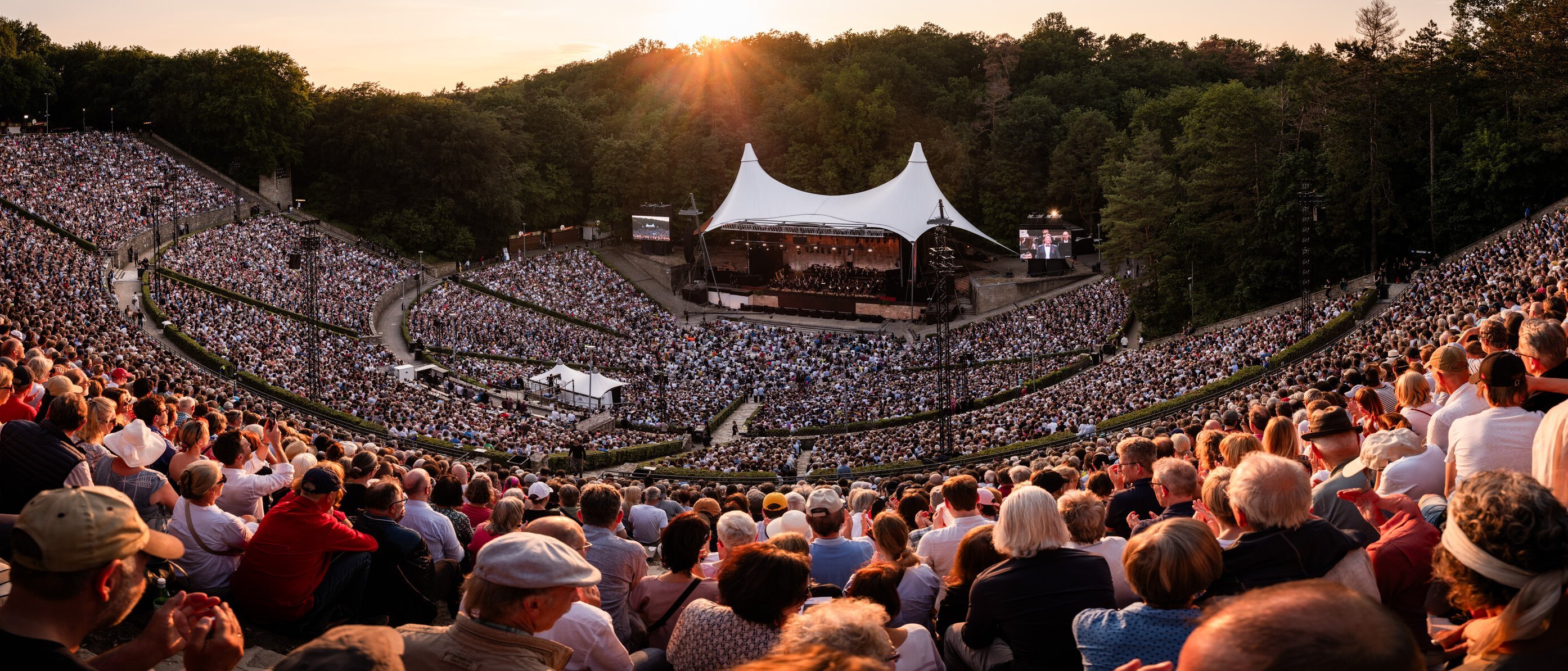 Ein großes Amphitheater unter freiem Himmel, vollgepackt mit Menschen, die ein Konzert bei Sonnenuntergang verfolgen. Die Bühne ist von einem weißen Baldachin bedeckt, umgeben von üppig grünen Bäumen unter einem warmen Himmel.