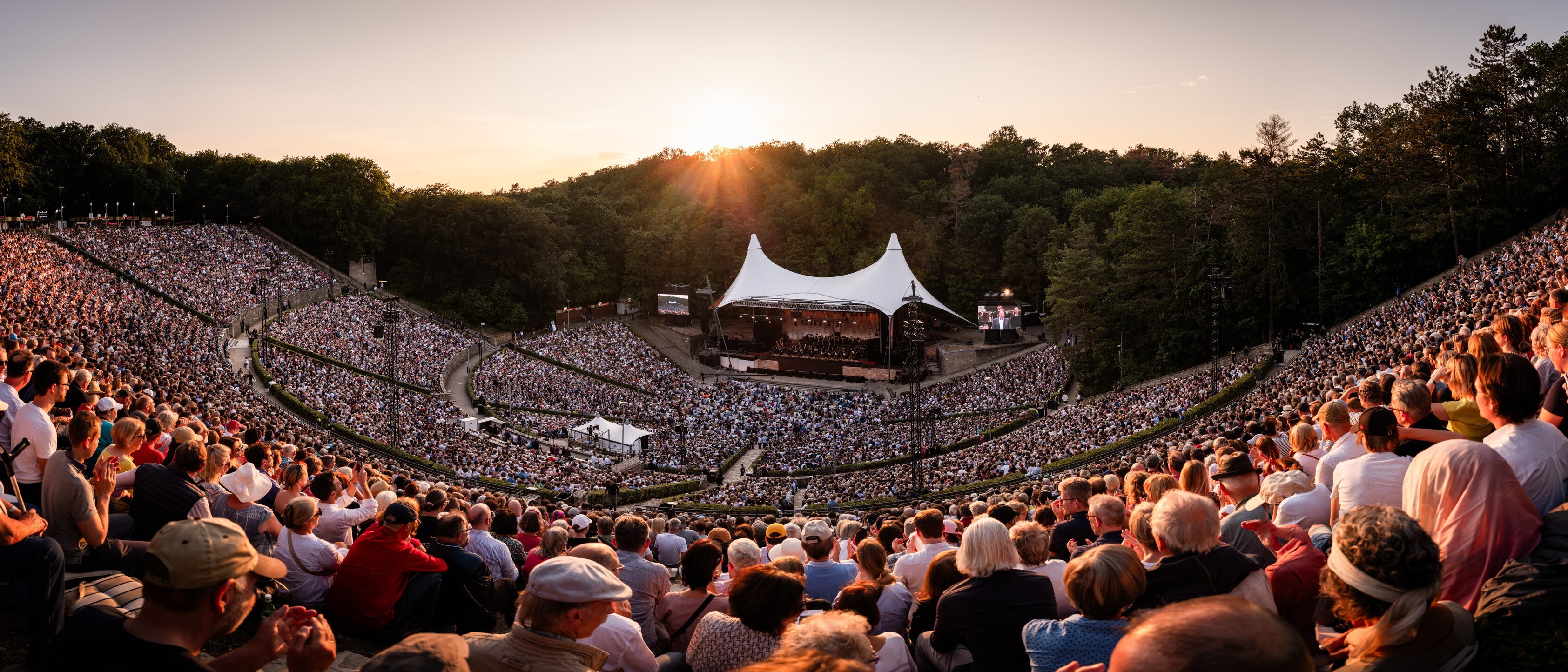 Ein großes Amphitheater unter freiem Himmel, vollgepackt mit Menschen, die ein Konzert bei Sonnenuntergang verfolgen. Die Bühne ist von einem weißen Baldachin bedeckt, umgeben von üppig grünen Bäumen unter einem warmen Himmel.