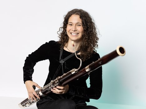 A smiling person with curly hair, wearing a black outfit, is sitting and holding a bassoon across their lap against a plain white background.