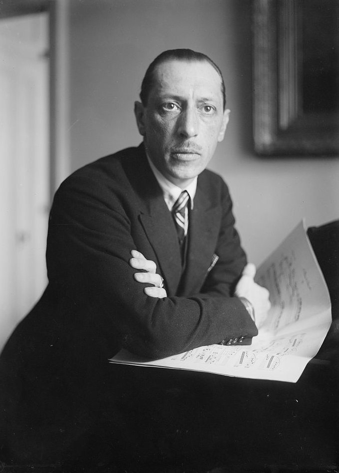 Igor Stravinsky, dressed in a suit, sits with his arms folded, holding sheets of music in his hand. He gazes directly into the camera with a serious expression. He is seated at a desk in a room with a framed picture on the wall behind him.so it reads like a refined portrait caption.