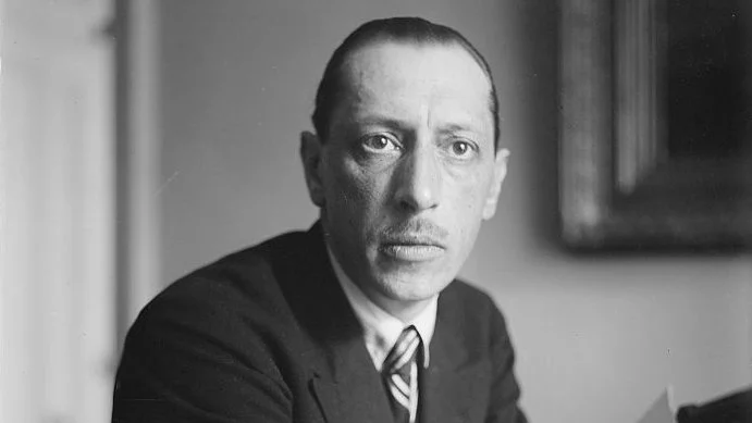 Igor Stravinsky, dressed in a suit, sits with his arms folded, holding sheets of music in his hand. He gazes directly into the camera with a serious expression. He is seated at a desk in a room with a framed picture on the wall behind him.so it reads like a refined portrait caption.
