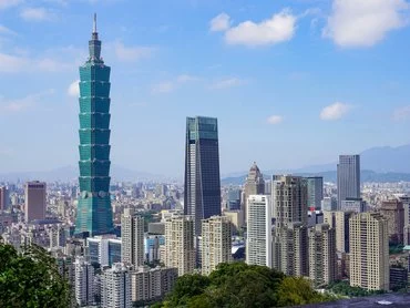 A cityscape view of Taipei with the tall, green Taipei 101 tower prominently rising above surrounding skyscrapers, framed by lush greenery and a clear, blue sky.